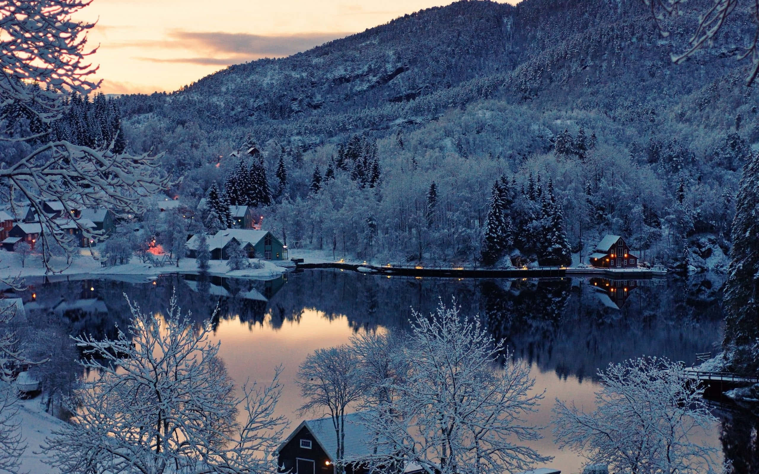 Snowy Village 桌布 壁紙 — 黃昏時分，白雪覆蓋的村莊依偎在平靜的湖邊，發光的窗戶倒映在冰冷的水面上。