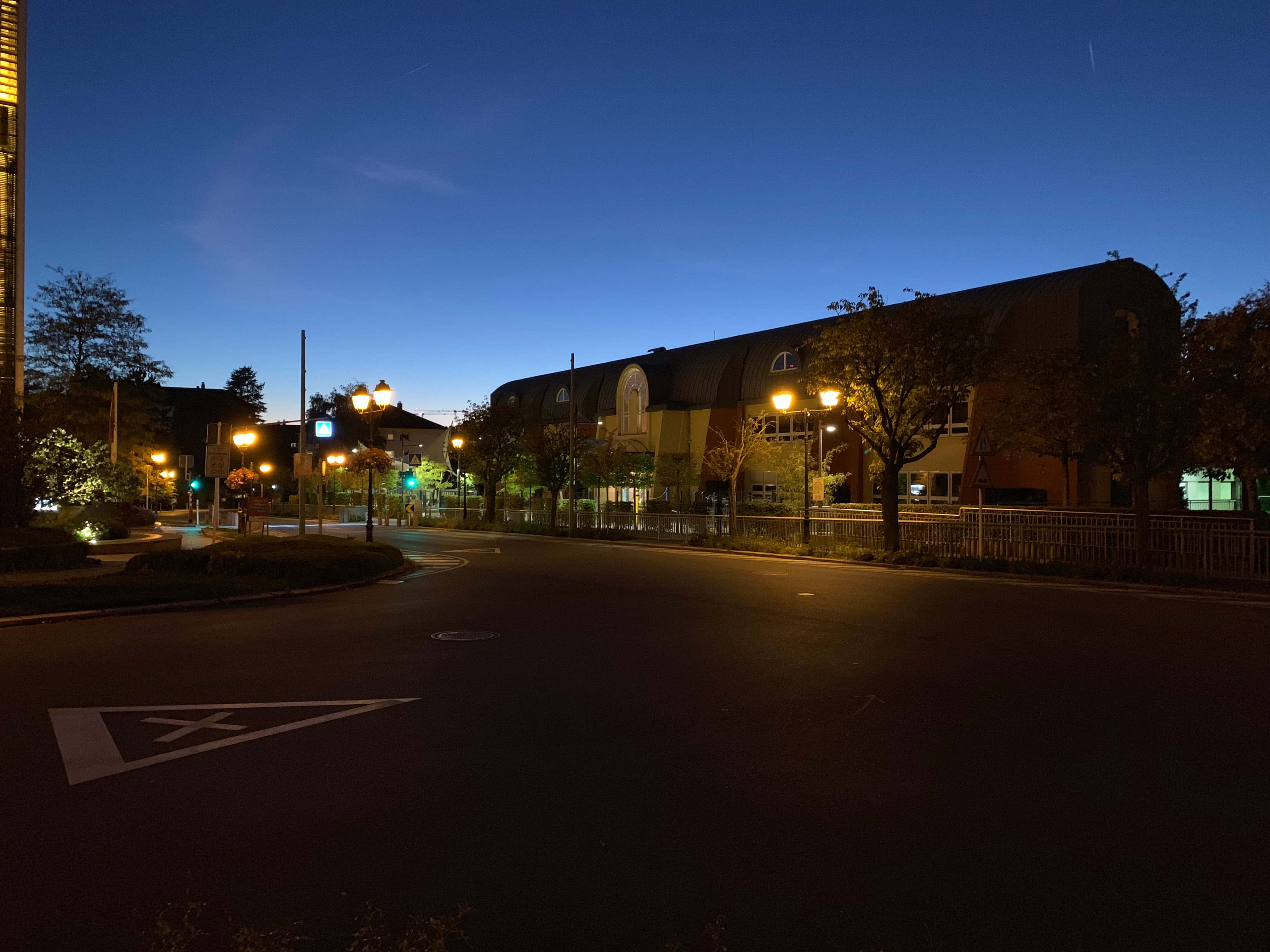 Night Mode 桌布 壁紙 — 黃昏時分一條寧靜的城市街道，溫暖的路燈照亮了一座弧形建築和樹木，襯托著深藍色的天空。