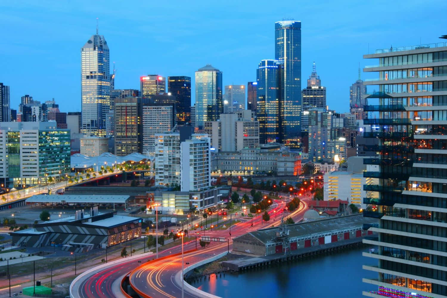 Adelaide 桌布 壁紙 — 黃昏時分充滿活力的城市天際線，閃爍的摩天大樓和蜿蜒河濱道路上紅色的車流燈光。