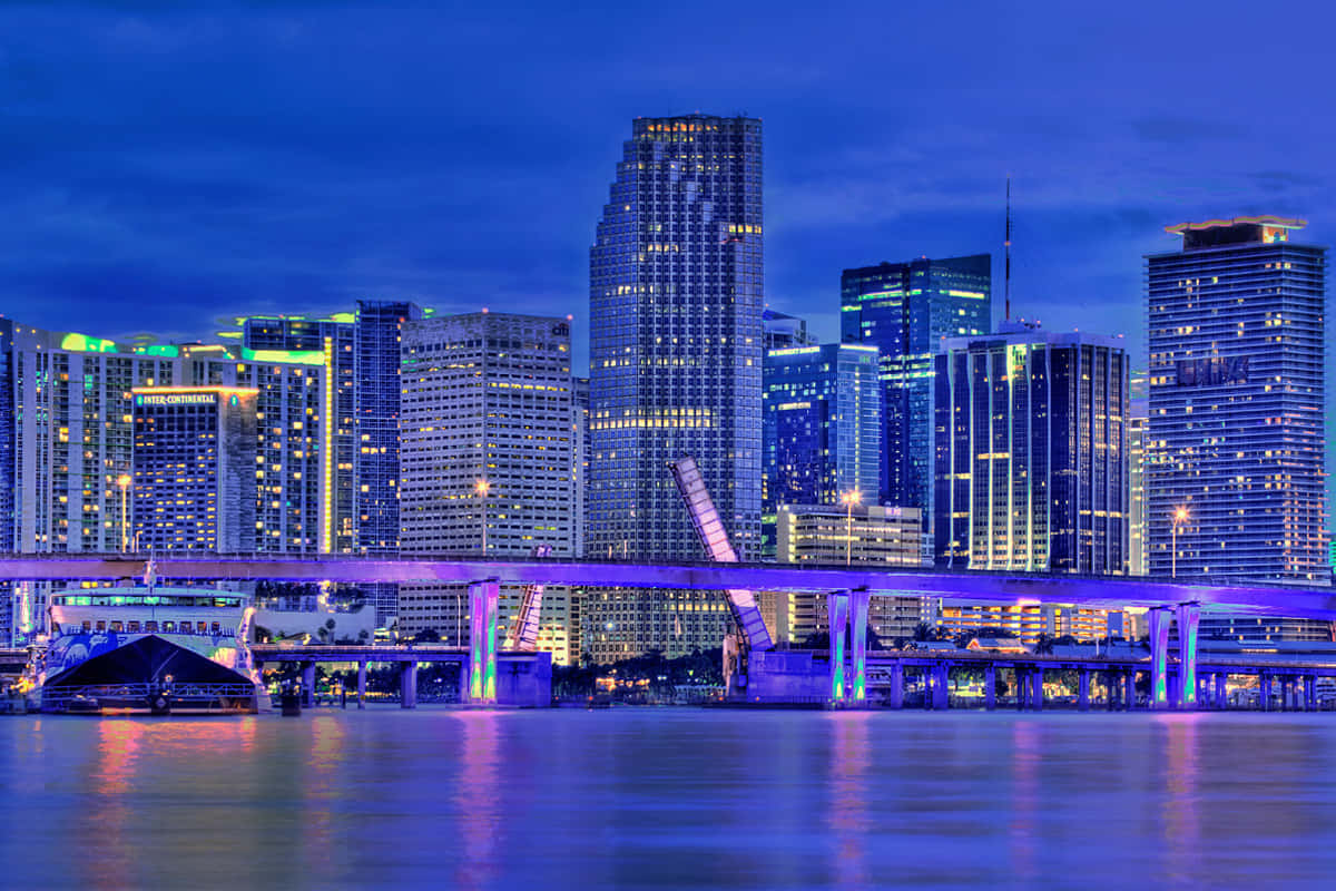 Miami 桌布 壁紙 — 黃昏時分充滿活力的城市天際線，閃爍的摩天大樓和紫色燈光的橋樑倒映在平靜的水面上。