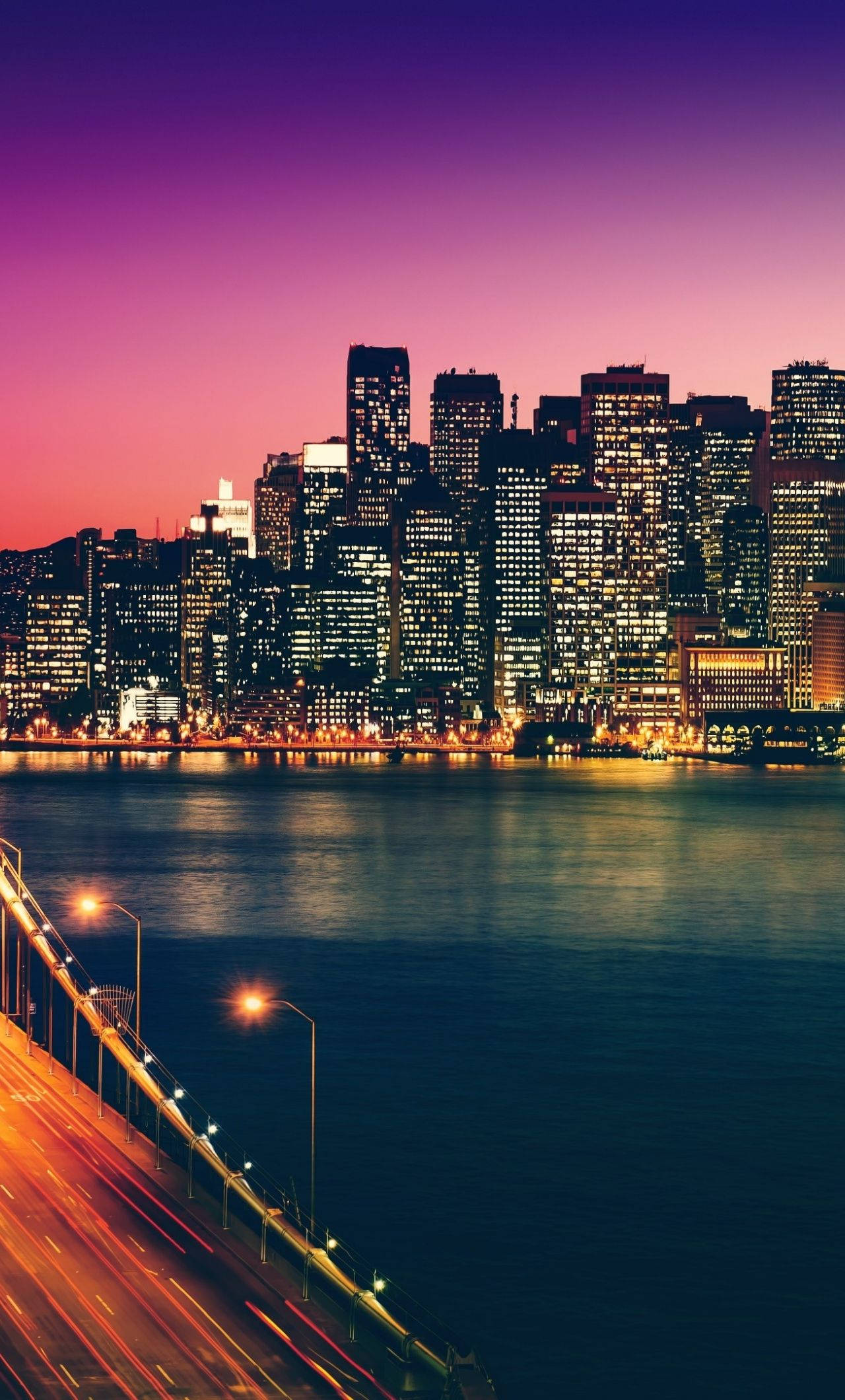San Francisco Phone 桌布 壁紙 — 黃昏時分充滿活力的城市天際線，閃爍的摩天大樓倒映在平靜的水面上，下方橋樑的光帶延伸開來。