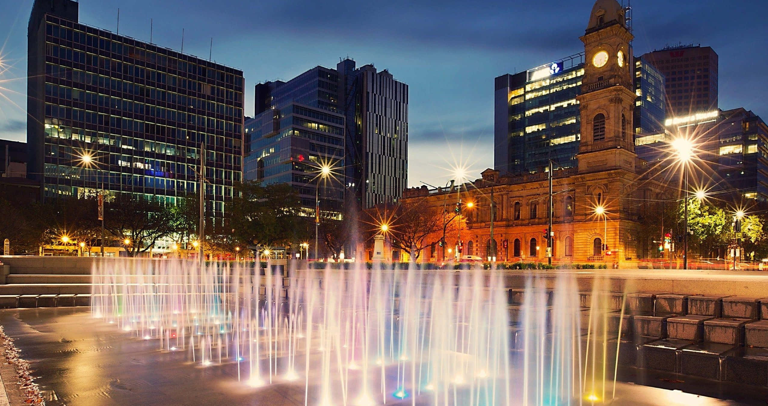 Adelaide 桌布 壁紙 — 黃昏時分充滿活力的城市廣場，噴泉在現代摩天大樓和歷史鐘樓的背景下噴射彩色水花。