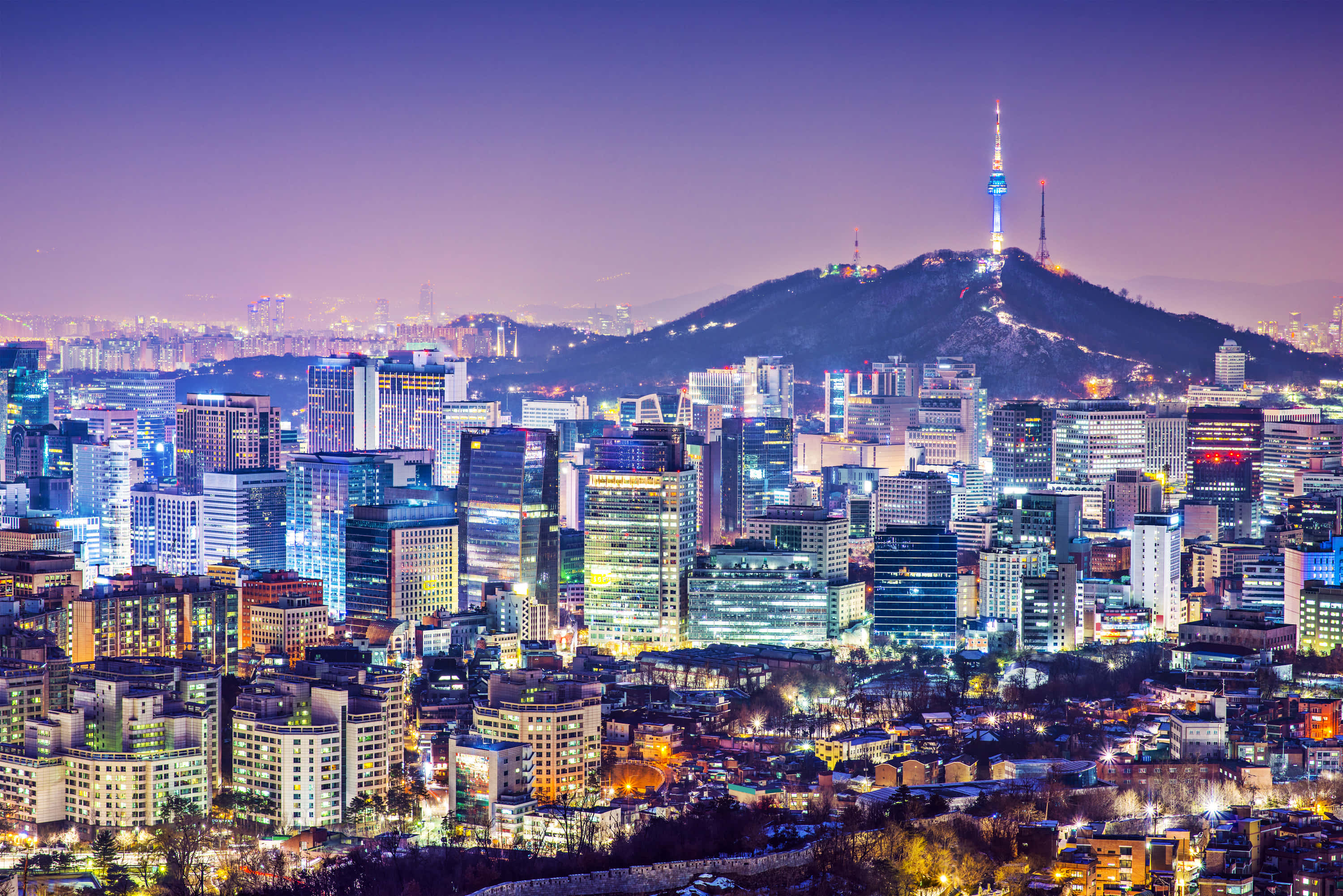 South Korea 桌布 壁紙 — 黃昏時分充滿活力的城市景觀，閃爍的摩天大樓和背景中一座頂部有燈塔的山脈。