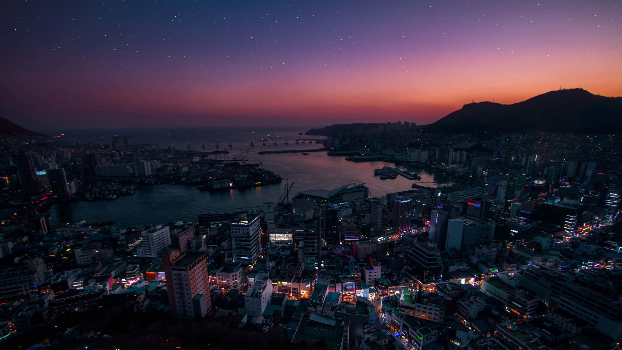 South Korea 桌布 壁紙 — 黃昏時分充滿活力的城市景觀，霓虹燈在紫色和橙色星空下的水面上閃爍倒映。