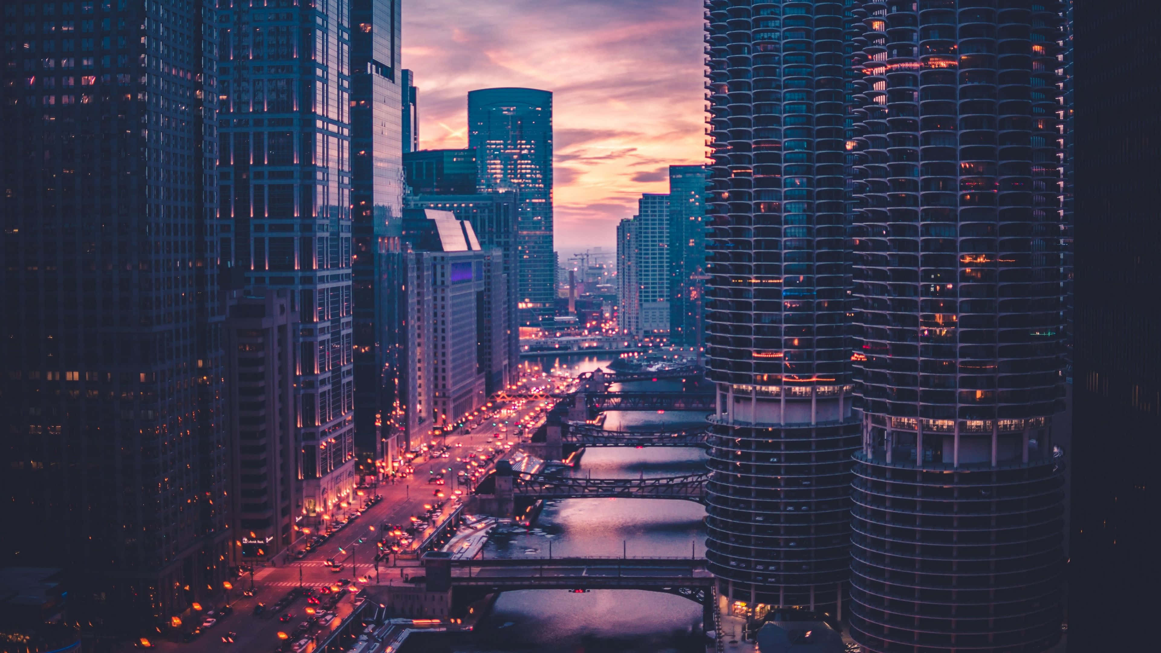 芝加哥 桌布 壁紙 — 黃昏時分充滿活力的城市景觀，高聳的摩天大樓在繁忙的河流上反射出粉紅色和紫色的天空，河流兩旁有橋樑和發光的街燈。