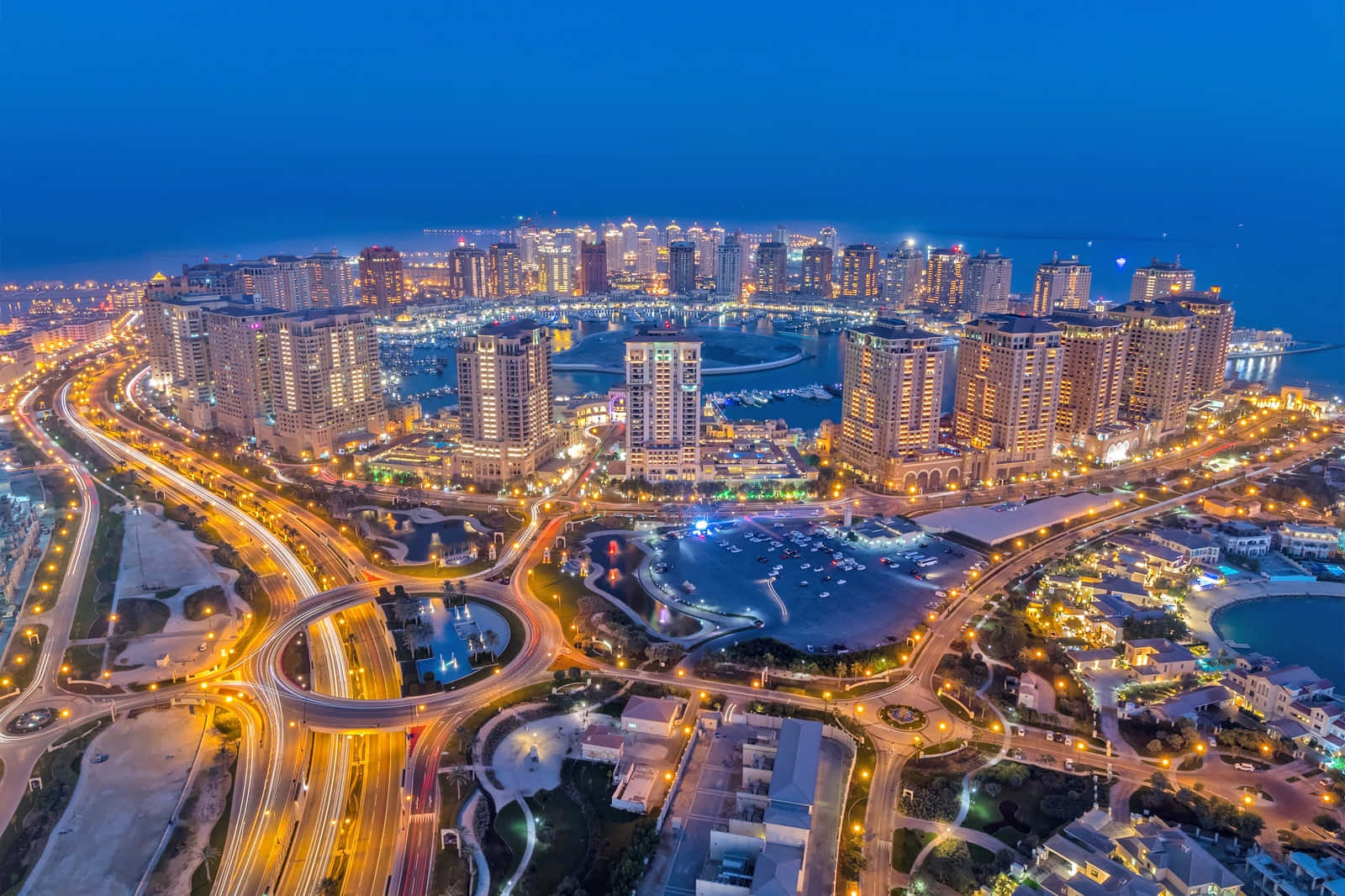 Qatar 桌布 壁紙 — 黃昏時分充滿活力的沿海城市的鳥瞰圖，有發光的摩天大樓、蜿蜒的道路和停滿船隻的碼頭。