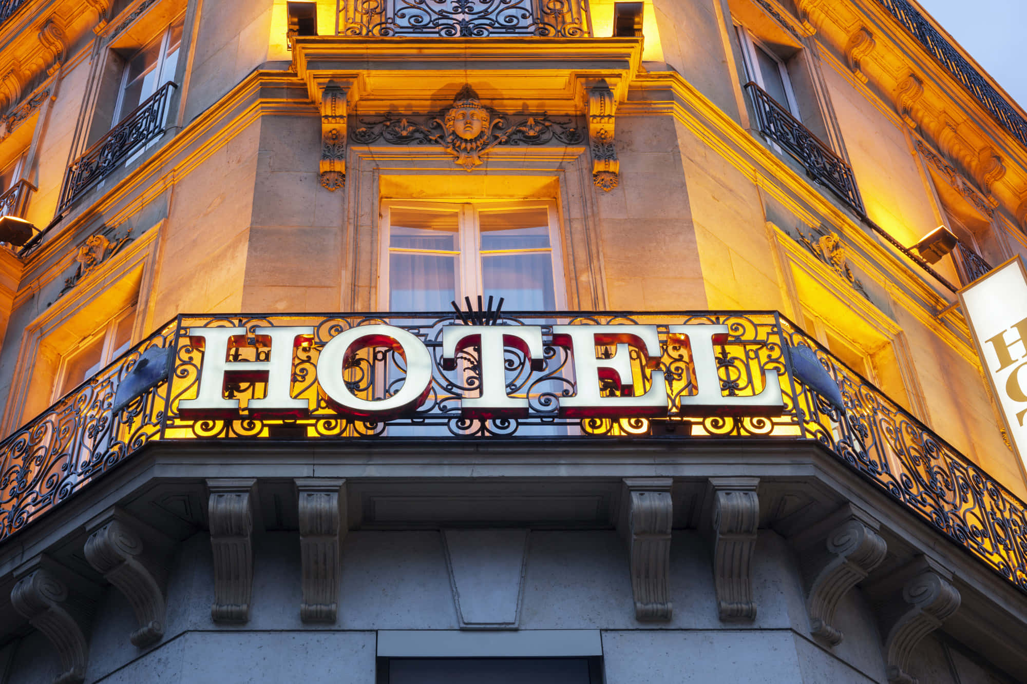 Hotel 桌布 壁紙 — 黃昏時分宏偉的酒店外牆，飾有華麗的鐵藝，發光的「HOTEL」招牌在石造建築上投下溫暖的金色光芒。
