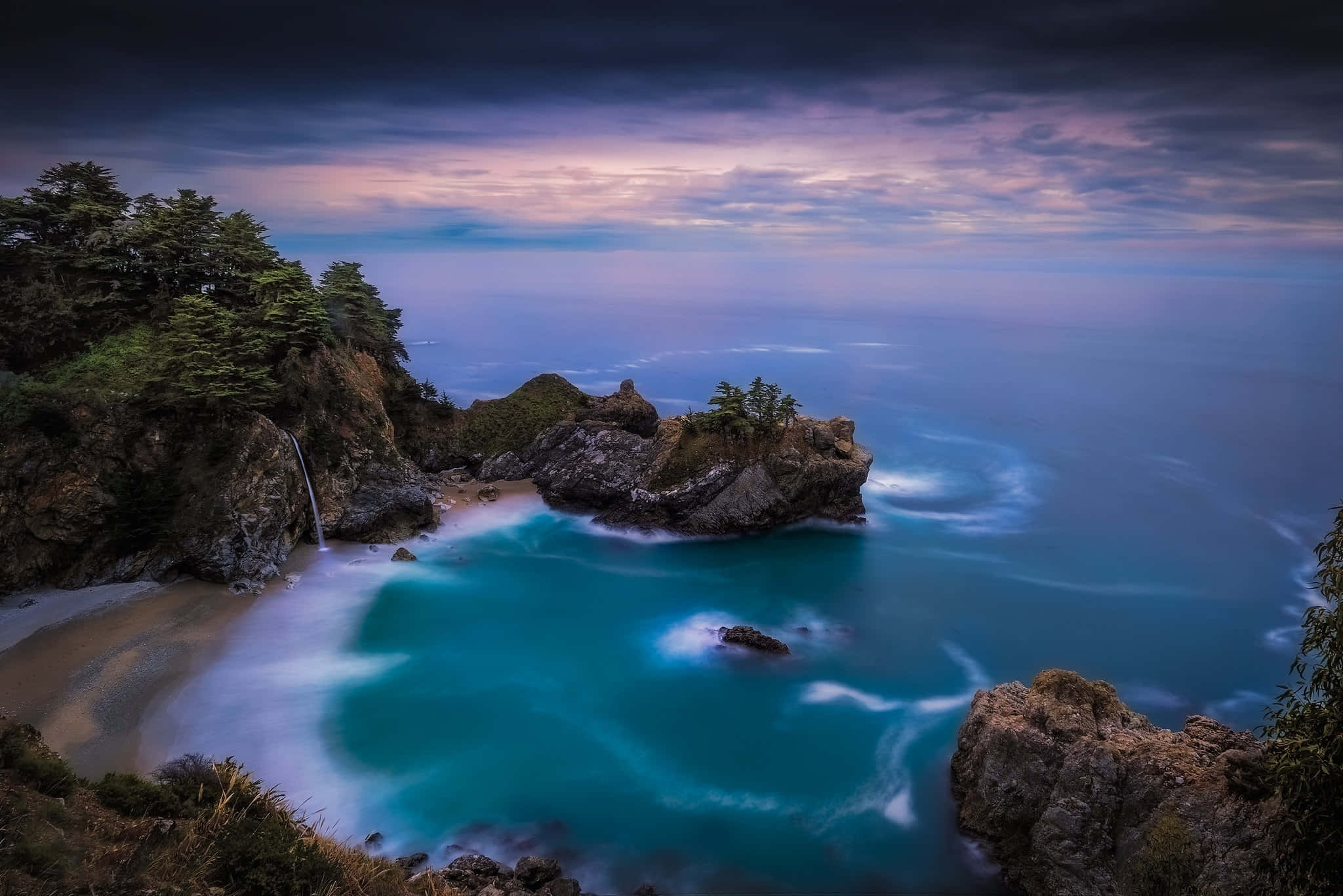 Big Sur 桌布 壁紙 — 黃昏時分寧靜的海岸小海灣，藍綠色的海水在岩石露頭周圍旋轉，瀑布傾瀉入海。