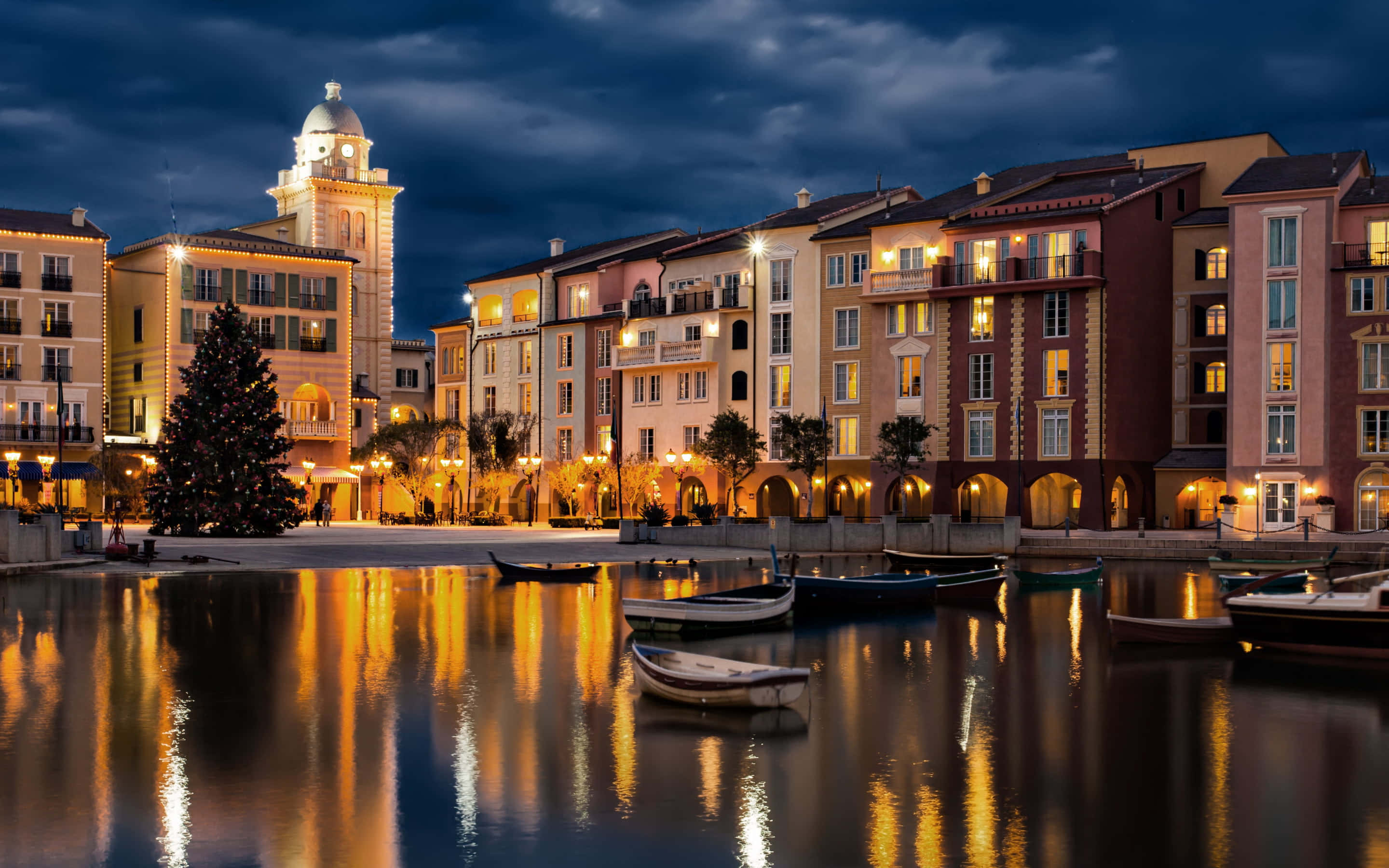 Orlando 桌布 壁紙 — 黃昏時分寧靜的海濱小鎮，溫暖燈光照亮的建築物和船隻在平靜的水面上反射出金色的光芒。