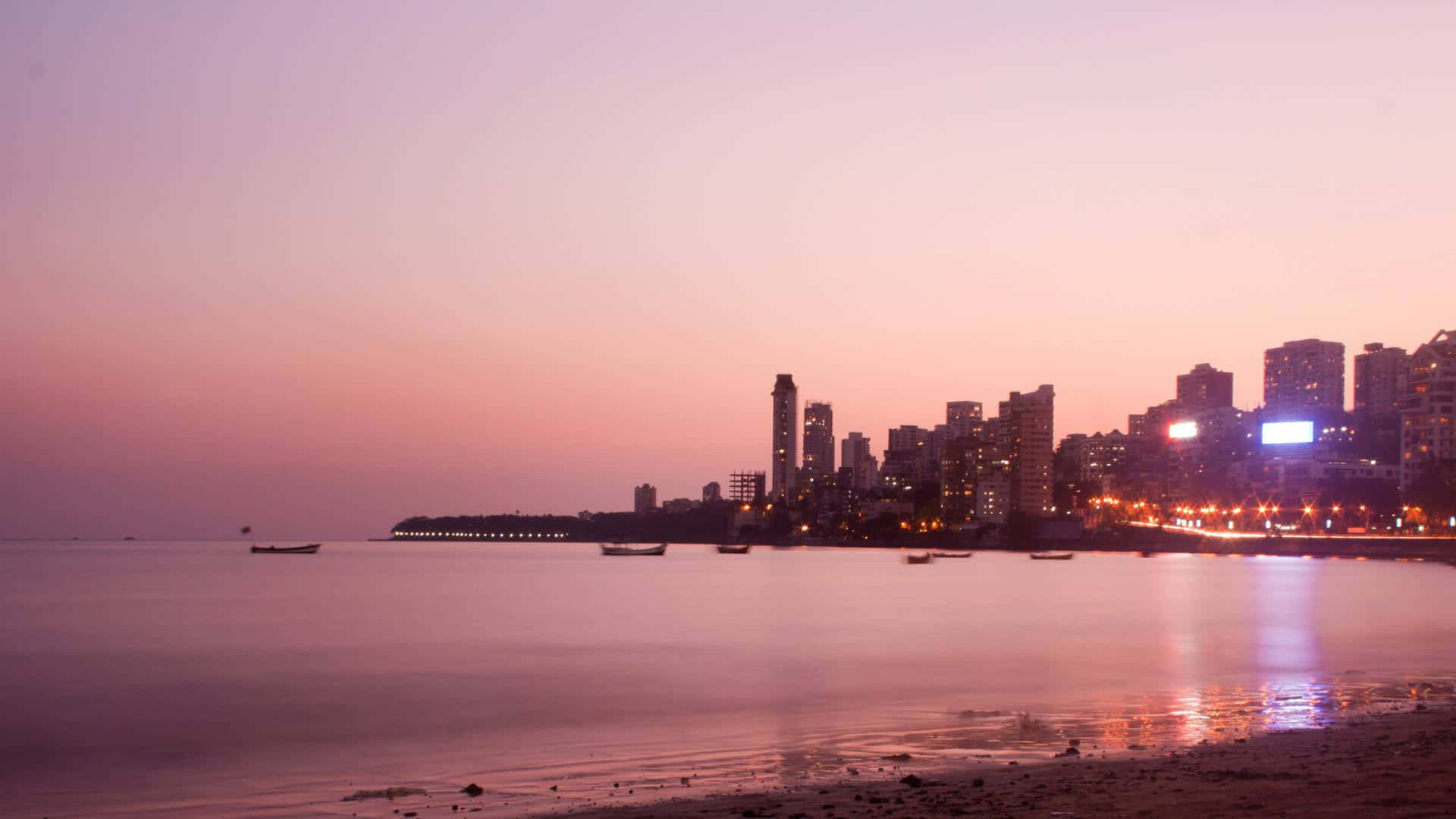 Mumbai 桌布 壁紙 — 黃昏時分寧靜的海灘，平靜的水面倒映著城市燈光和船隻的剪影，襯托著柔和的粉紅色和紫色天空。
