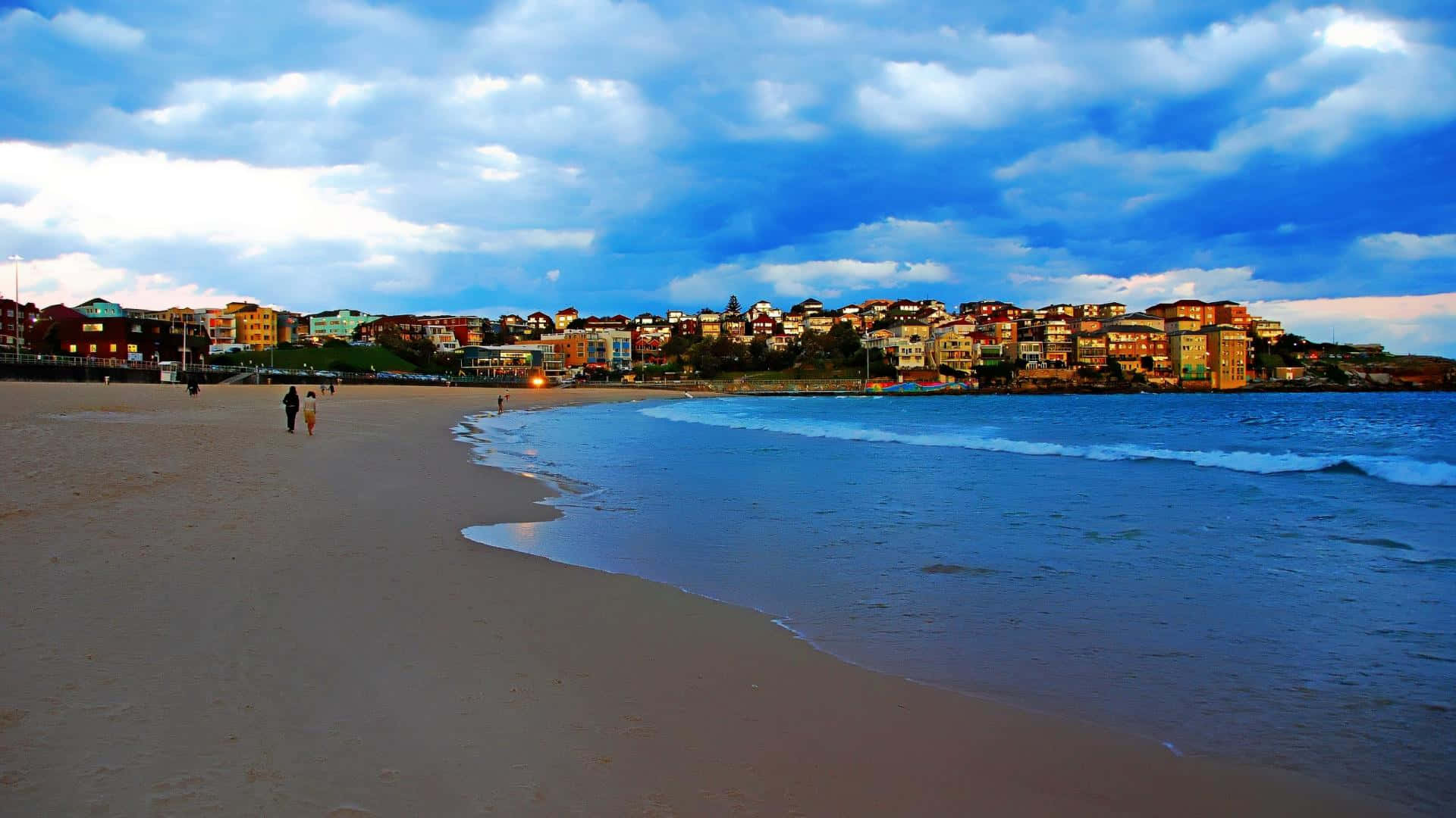 Australia Beach 桌布 壁紙 — 黃昏時分寧靜的海灘，溫柔的波浪輕拍沙灘，遠處色彩繽紛的房屋在戲劇性的藍天下排列。