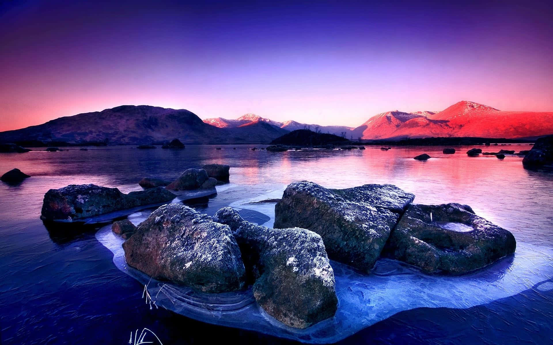 Frozen Lake 桌布 壁紙 — 黃昏時分寧靜的湖景，白雪覆蓋的山脈倒映在深藍色的水中，四周環繞著巨石。