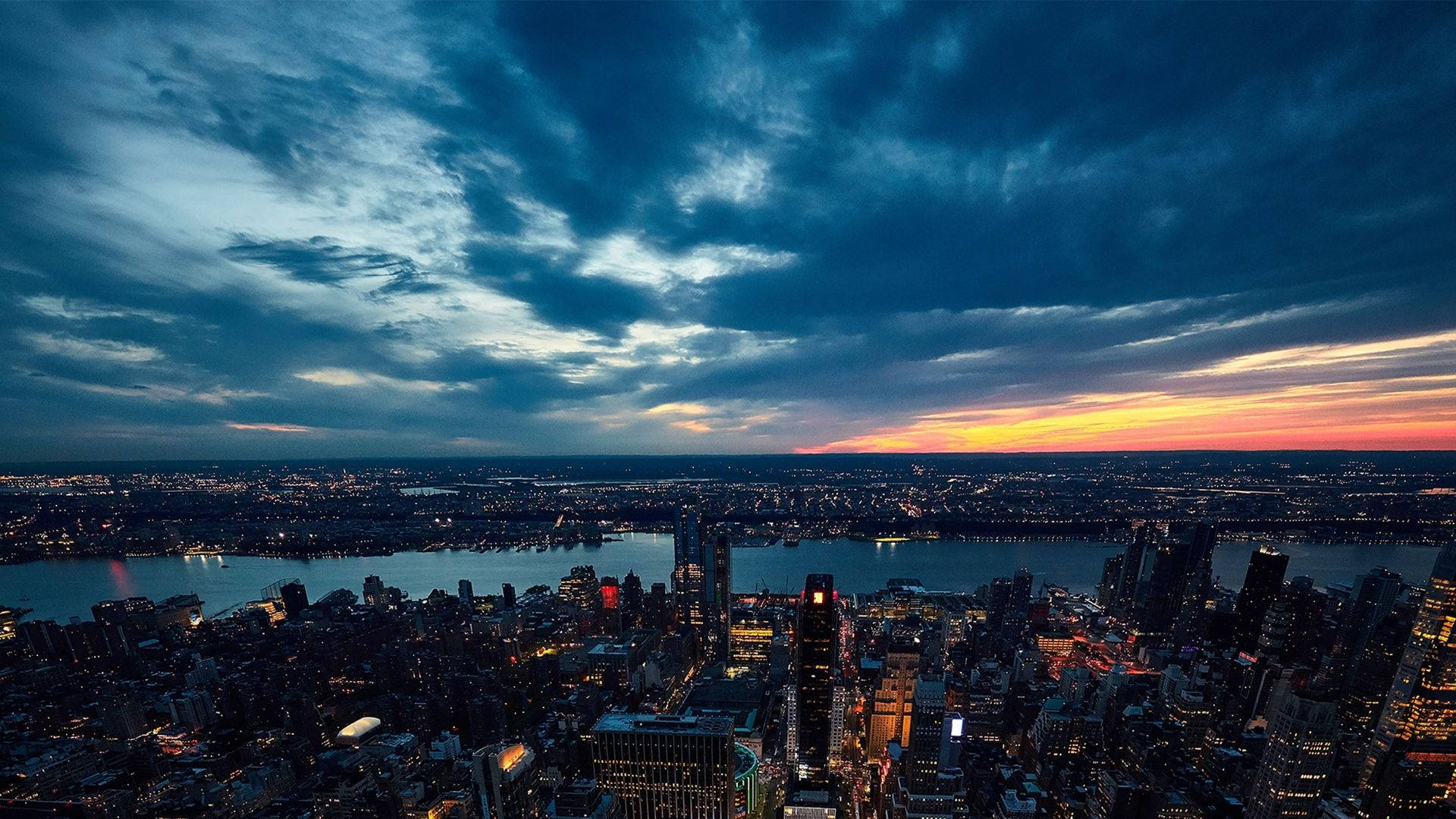 View 桌布 壁紙 — 黃昏時分廣闊的城市景觀，閃爍的摩天大樓和戲劇性雲層下充滿活力的橙色地平線。