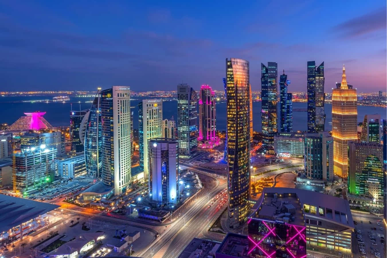 Qatar 桌布 壁紙 — 黃昏時分未來城市天際線的鳥瞰圖，霓虹燈照亮的高樓大廈倒映在水面上，道路閃爍著光芒。