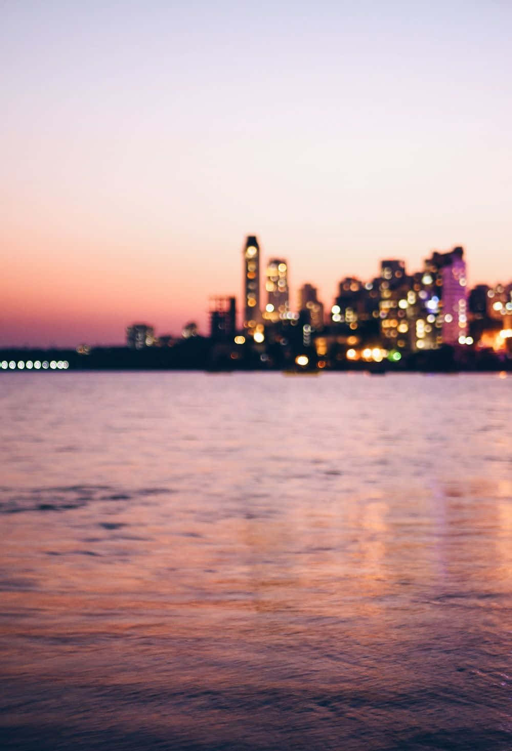 Mumbai 桌布 壁紙 — 黃昏時分模糊的城市天際線在平靜的水面上反射出溫暖的粉紅色和橙色燈光。