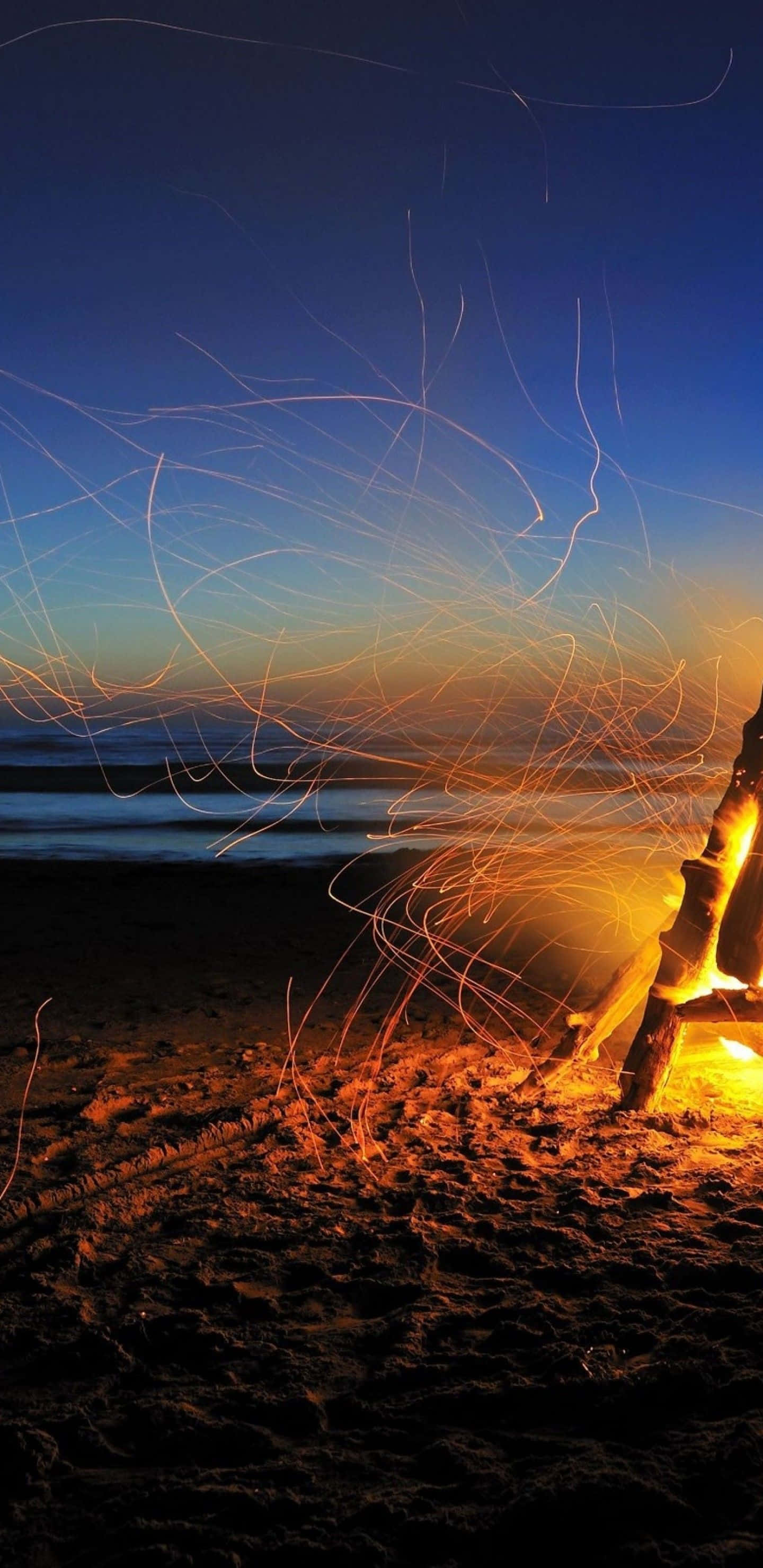 Beach Camping 桌布 壁紙 — 黃昏時分海灘上的營火，發光的橙色火花在深藍色天空和寧靜的海浪中舞動。