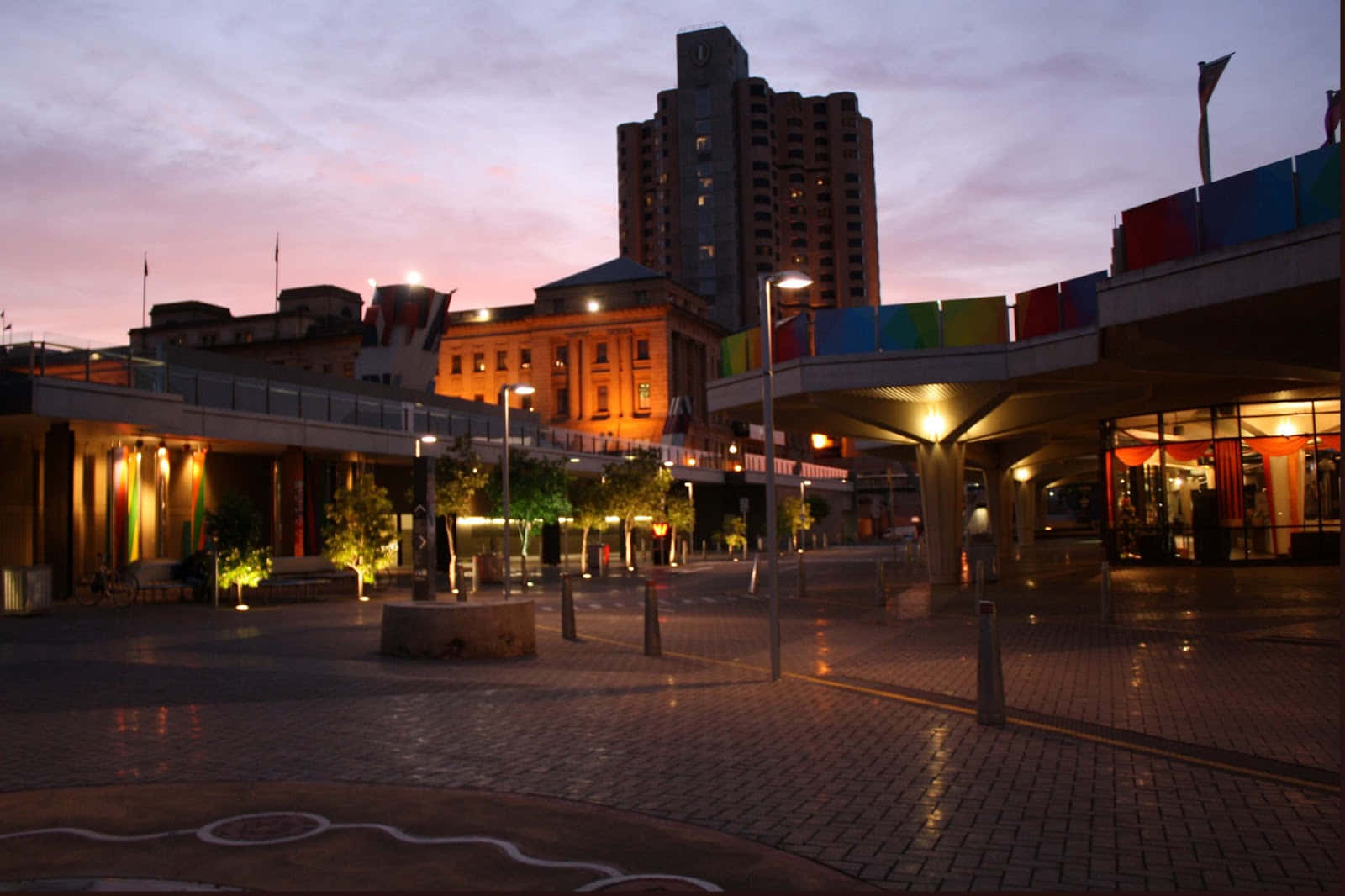 Adelaide 桌布 壁紙 — 黃昏時分濕潤的城市廣場，溫暖的燈光反射在路面上，色彩繽紛的旗幟襯托著紫色的天空。