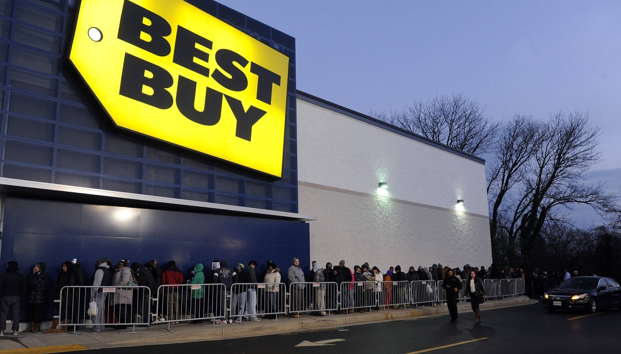 黃昏時分的Best Buy 壁紙 — 黃昏時分，人群在發光的黃色招牌下，以光禿樹木為背景，在Best Buy商店外排隊。