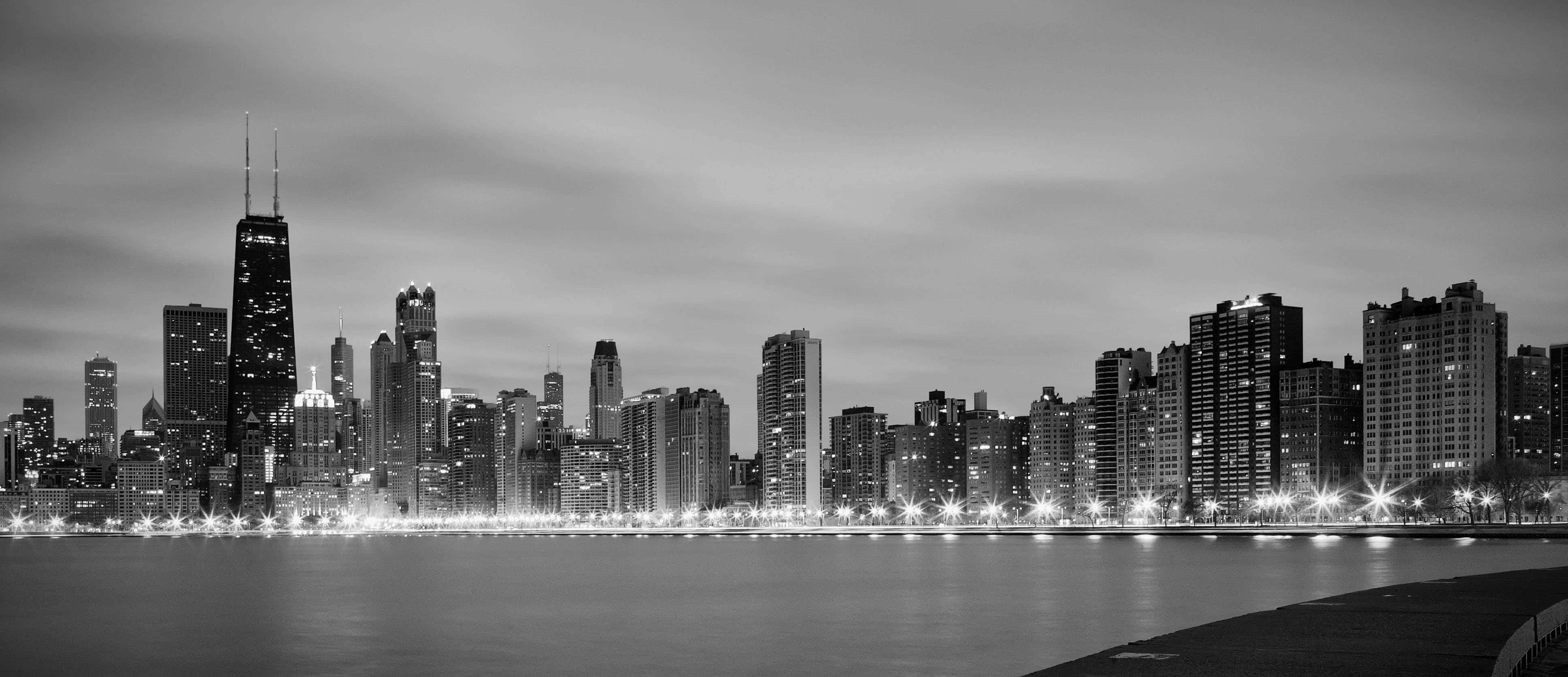 Black And White Cityscape 桌布 壁紙 — 黃昏時分的單色城市天際線，燈火通明的摩天大樓倒映在平靜的水面上。