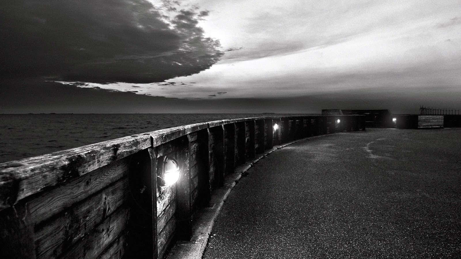 Dark Landscape 桌布 壁紙 — 黃昏時分的單色碼頭，發光的燈籠照亮風化的木頭和瀝青，天空多雲而戲劇化。