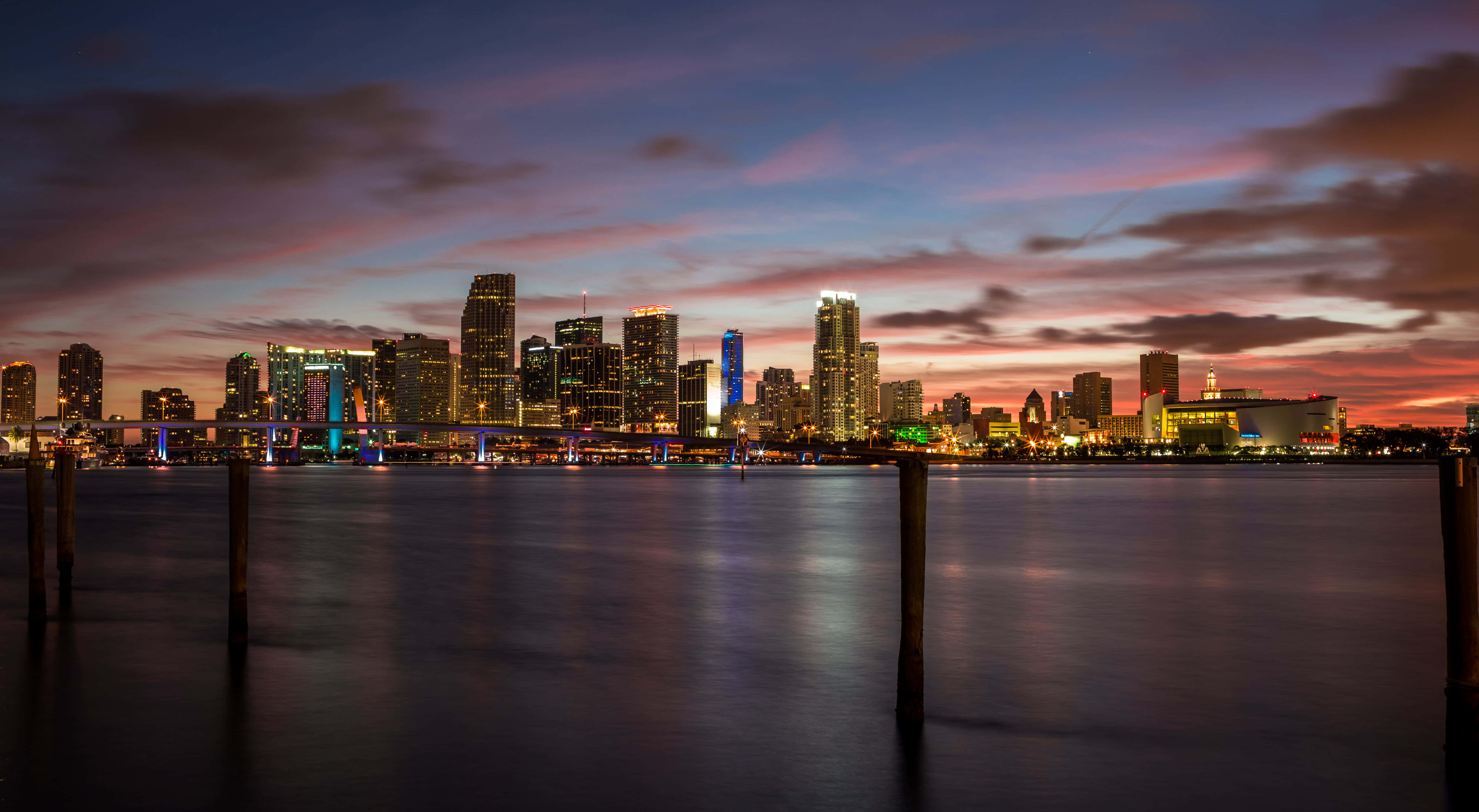 Miami 桌布 壁紙 — 黃昏時分的城市天際線，在紫色和橙色的天空下，發光的摩天大樓倒映在平靜的水面上。