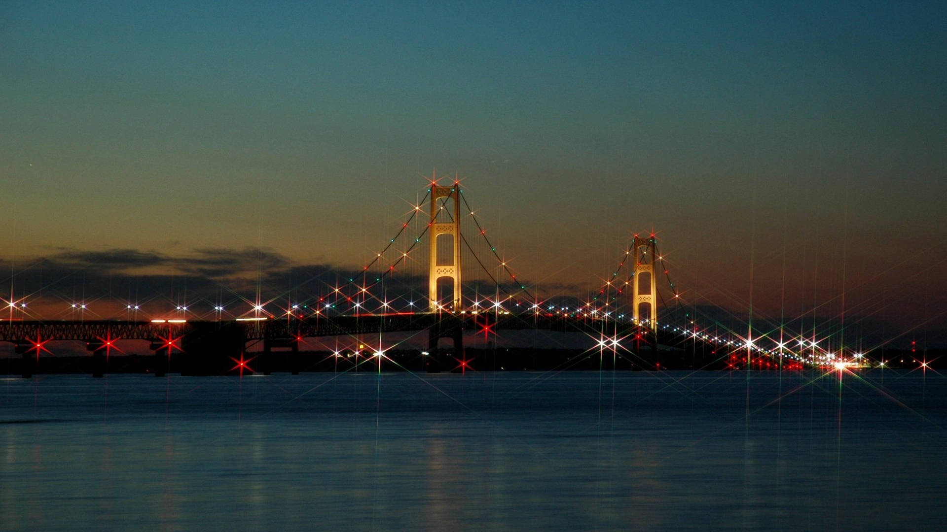 黃昏時分的壯麗麥基那克橋 壁紙 — 一座雄偉的吊橋在暮色中閃爍著節日燈光，映照在平靜的水面上，並帶有星爆效果。