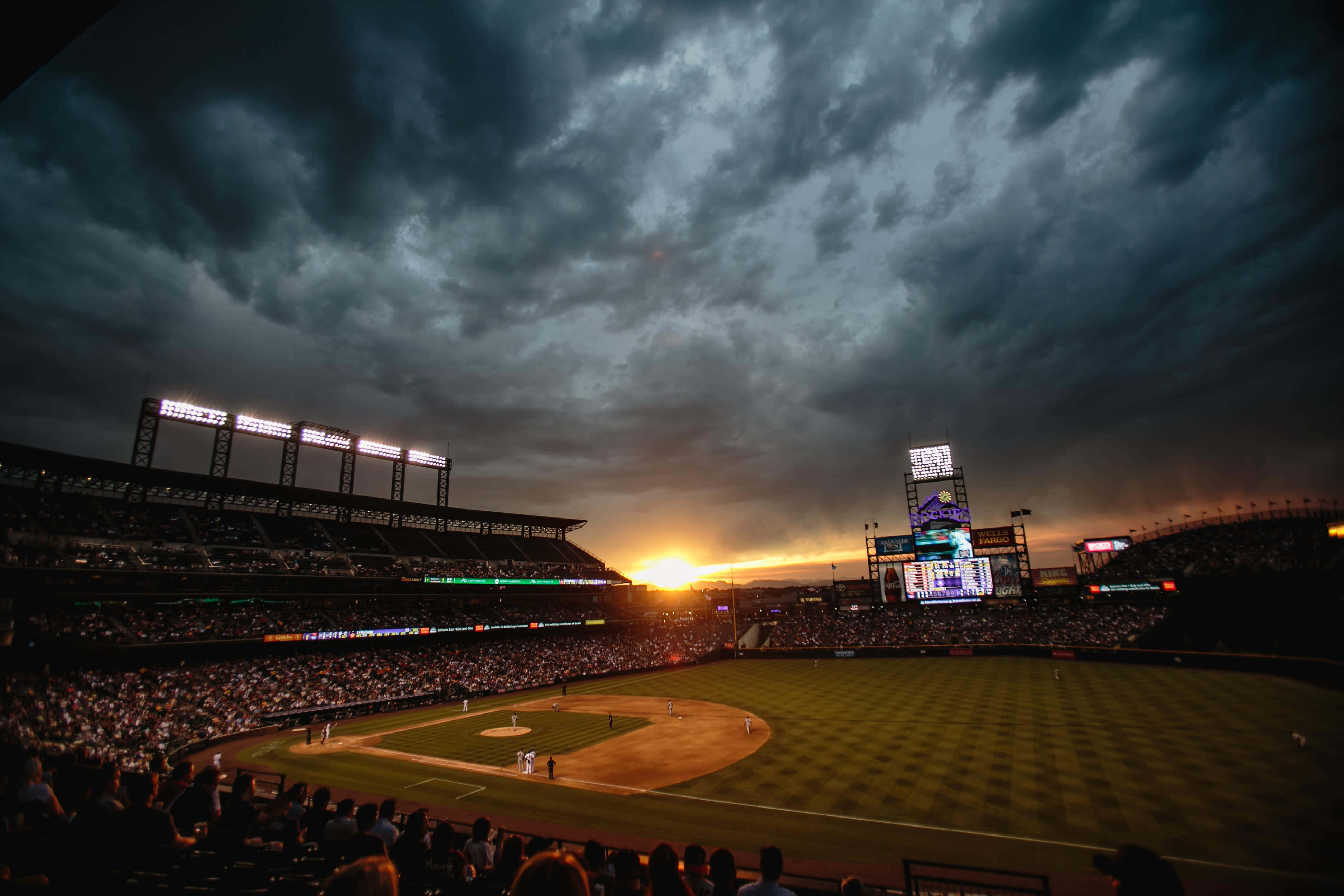 Baseball Stadium 桌布 壁紙 — 黃昏時分的棒球場，戲劇性的多雲天空和發光的場地燈光照亮了球場和人群。
