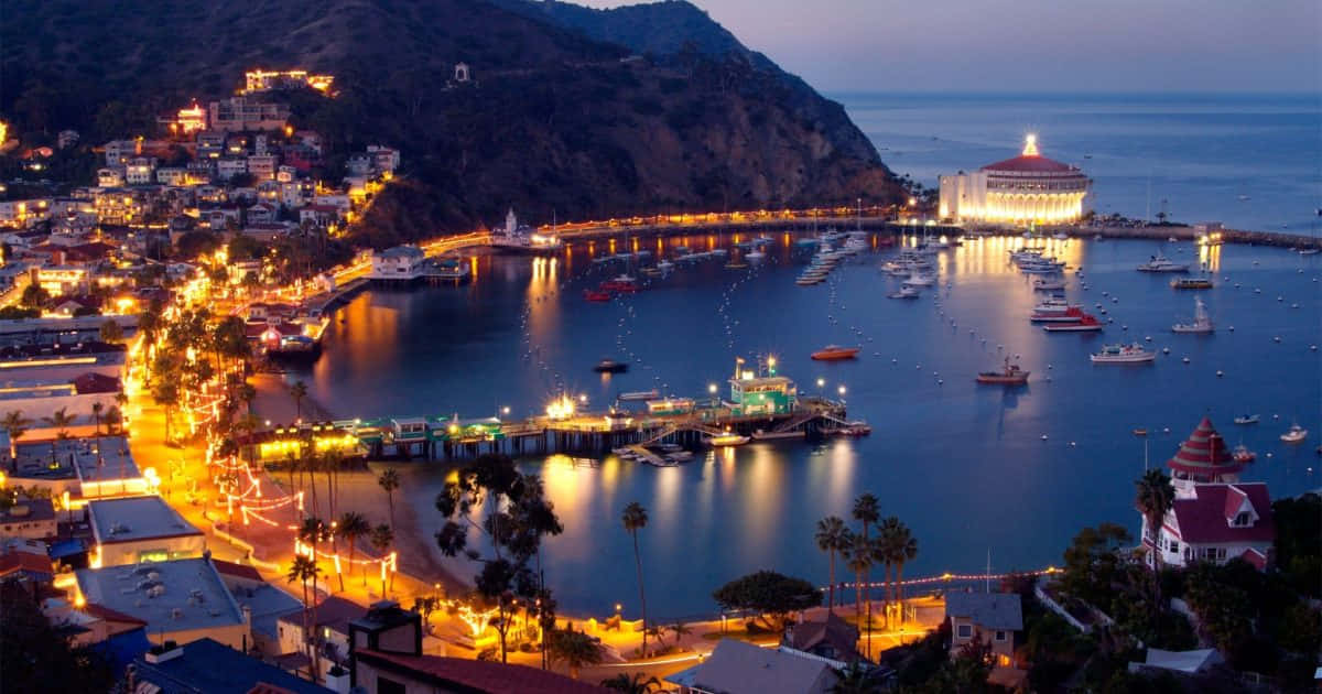 California Island 桌布 壁紙 — 黃昏時分的沿海小鎮，發光的街燈倒映在平靜的水面上，船隻停泊在一座宏偉的照明建築附近。