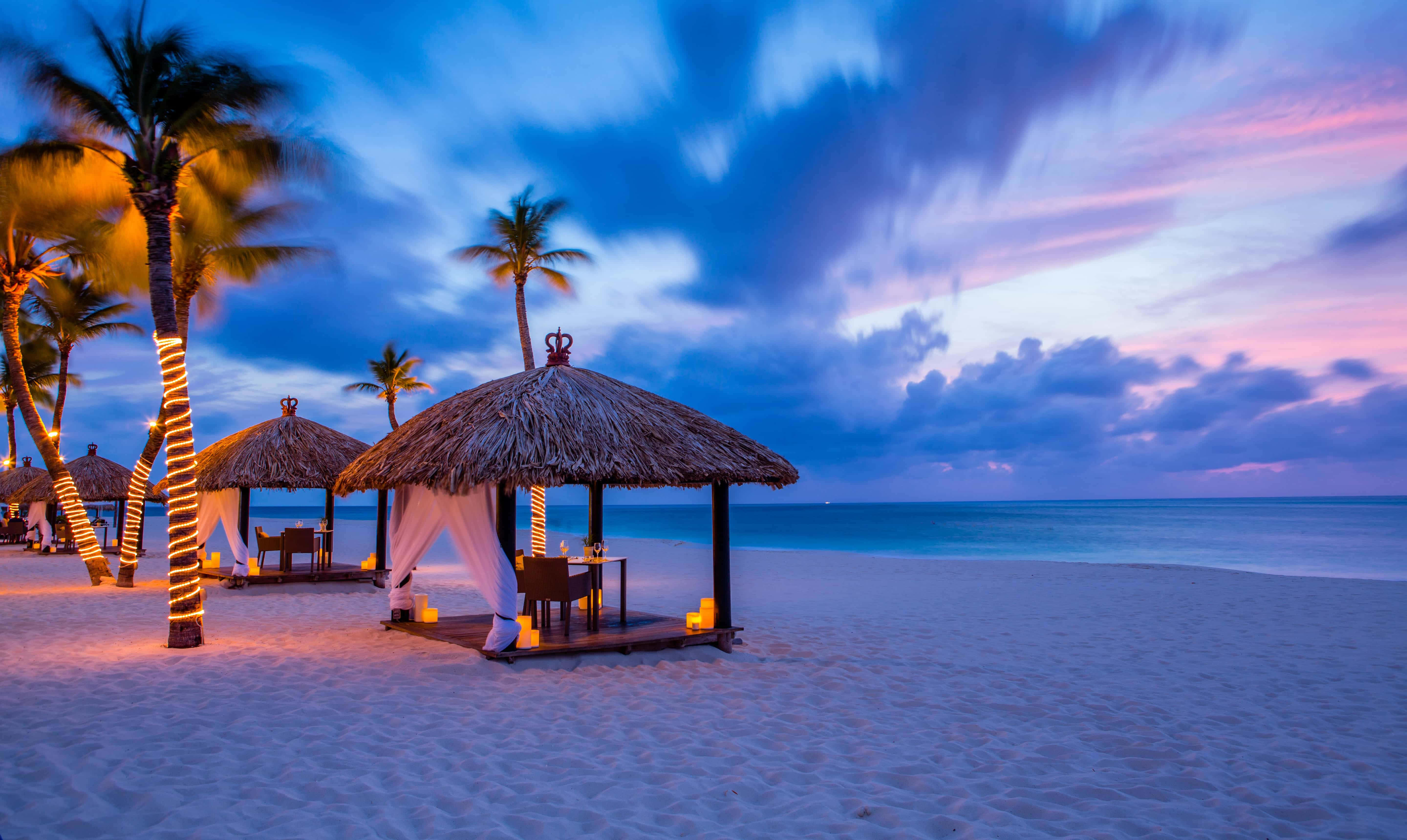 Aruba 桌布 壁紙 — 黃昏時分的熱帶海灘，茅草遮陽傘在充滿活力的紫色和粉紅色天空下溫暖地發光。
