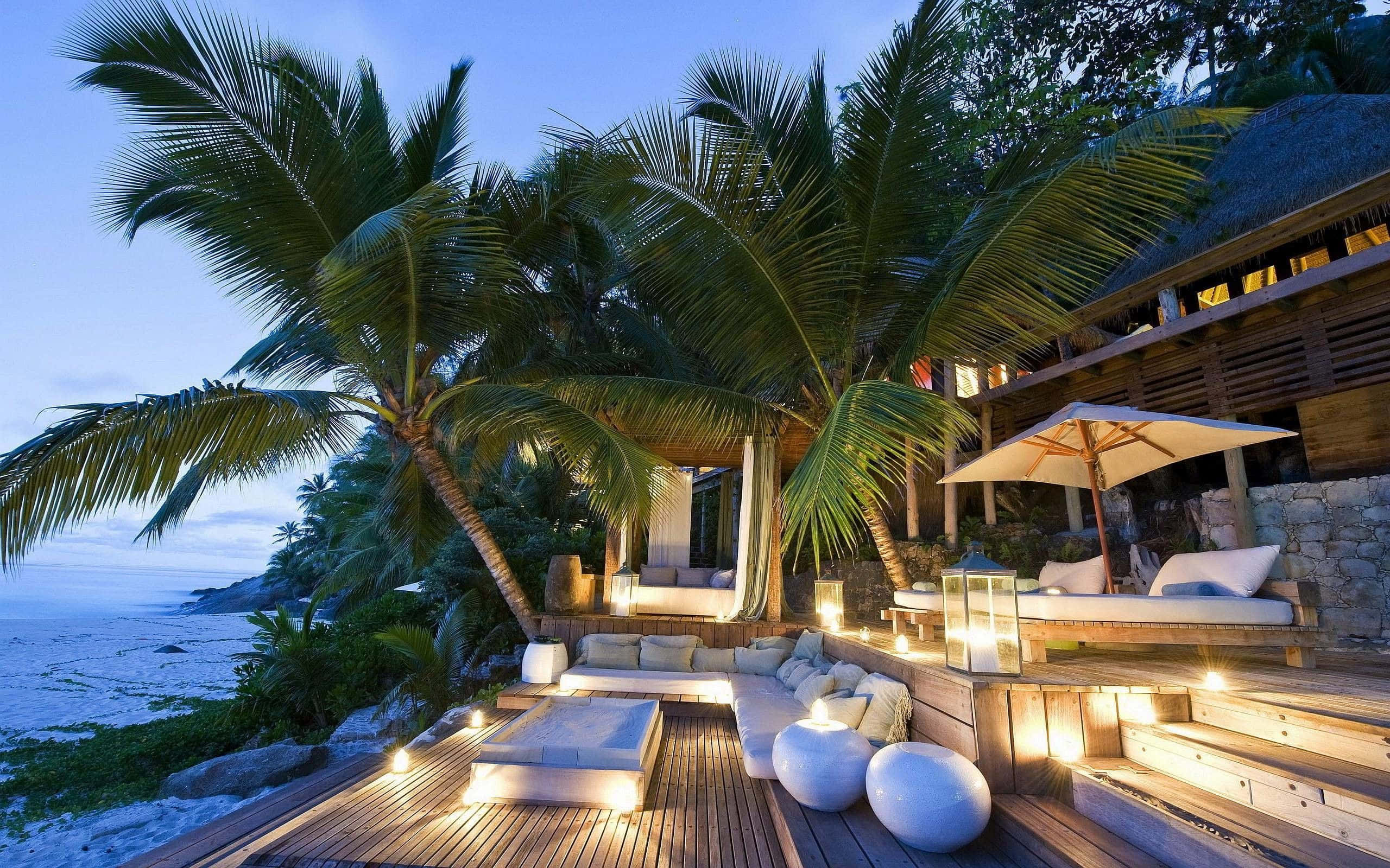 Beach Resort 桌布 壁紙 — 黃昏時分的豪華海灘甲板，棕櫚樹、發光燈籠和海洋景觀。
