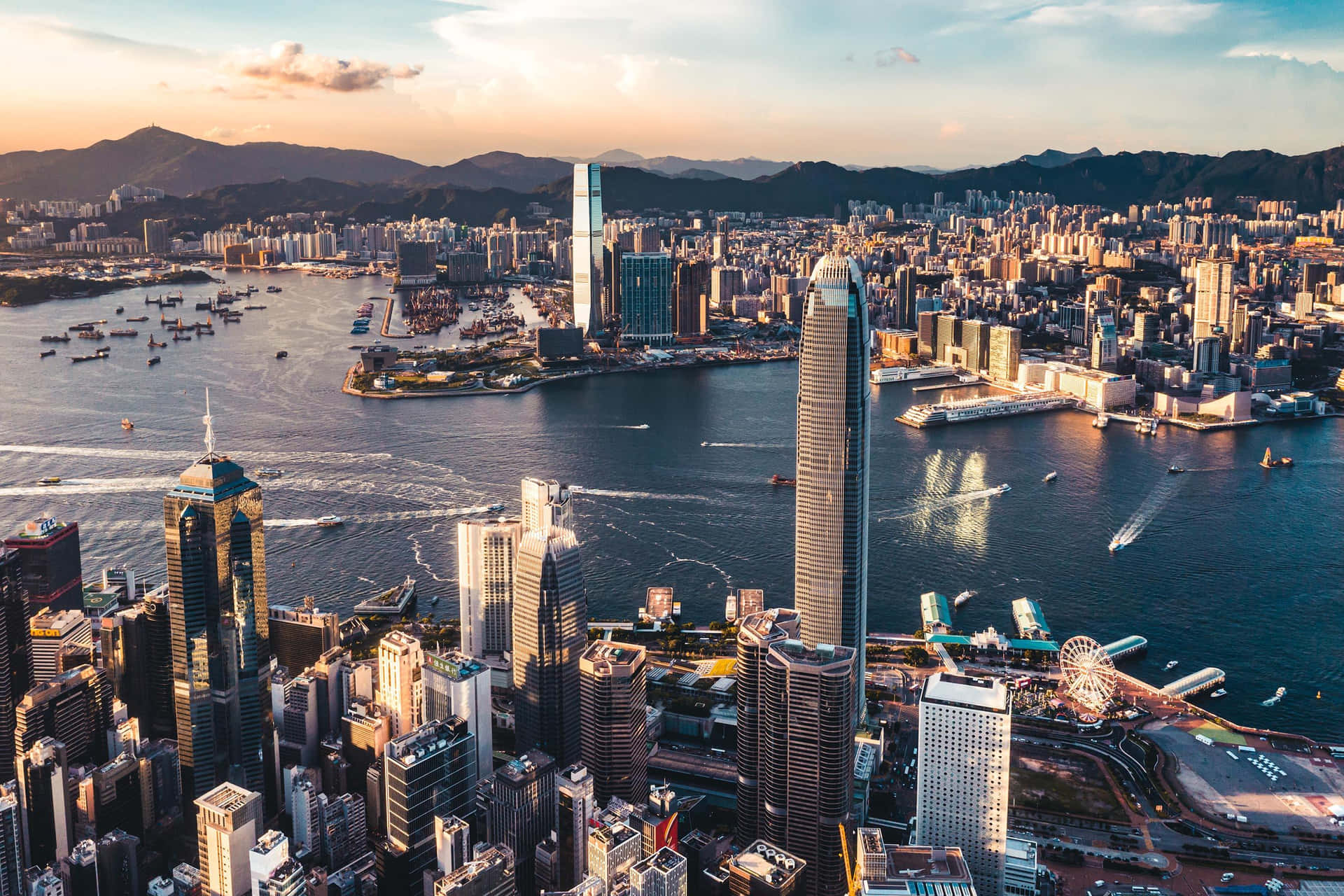 黃昏時分的香港天際線 壁紙 — 黃金時段香港天際線的鳥瞰圖，摩天大樓將溫暖的光線反射到充滿船隻的維多利亞港上。