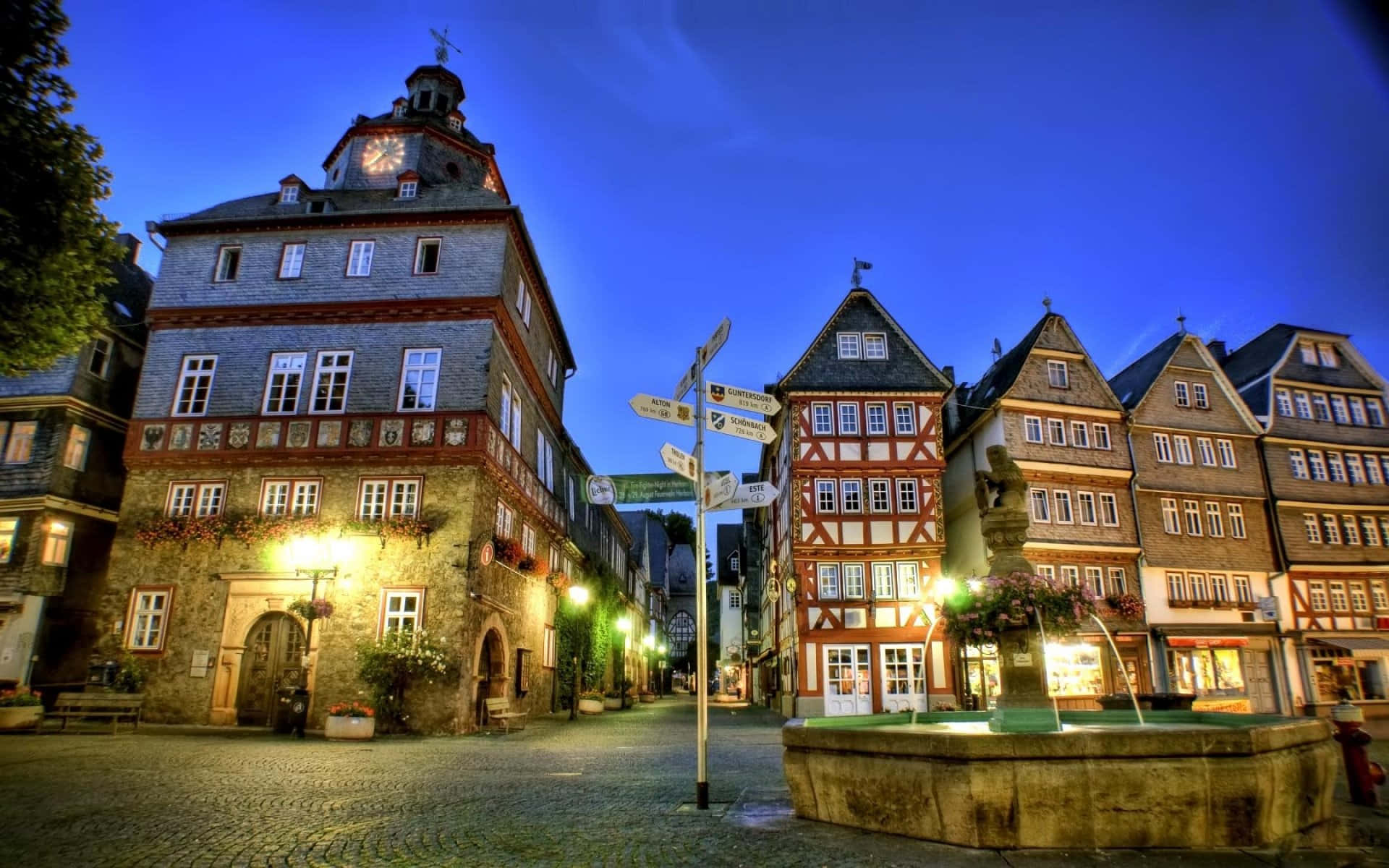 Germany 桌布 壁紙 — 黃昏時分的鵝卵石城鎮廣場，半木結構建築在深藍色天空下閃爍著溫暖的燈光。