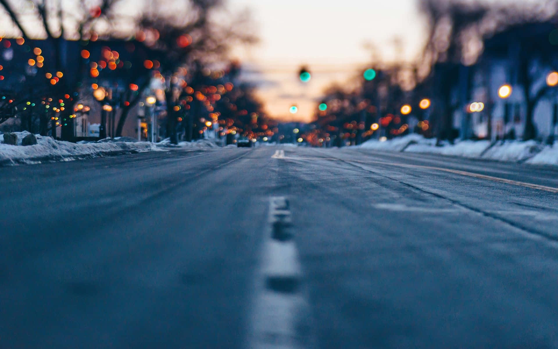 Icy Road 桌布 壁紙 — 黃昏時分積雪的都市街道，模糊的溫暖燈光與光禿的樹木排列在路旁。