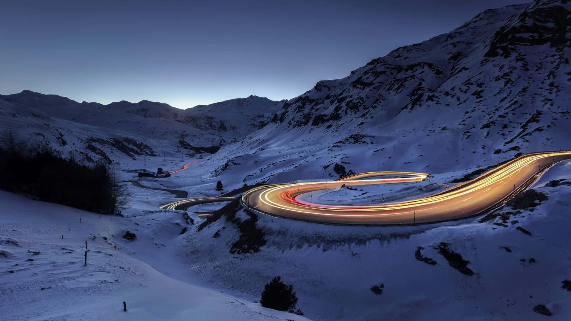 Snowy Road 桌布 壁紙 — 黃昏時分蜿蜒的山路，汽車發出的光芒穿過白雪覆蓋的山峰。