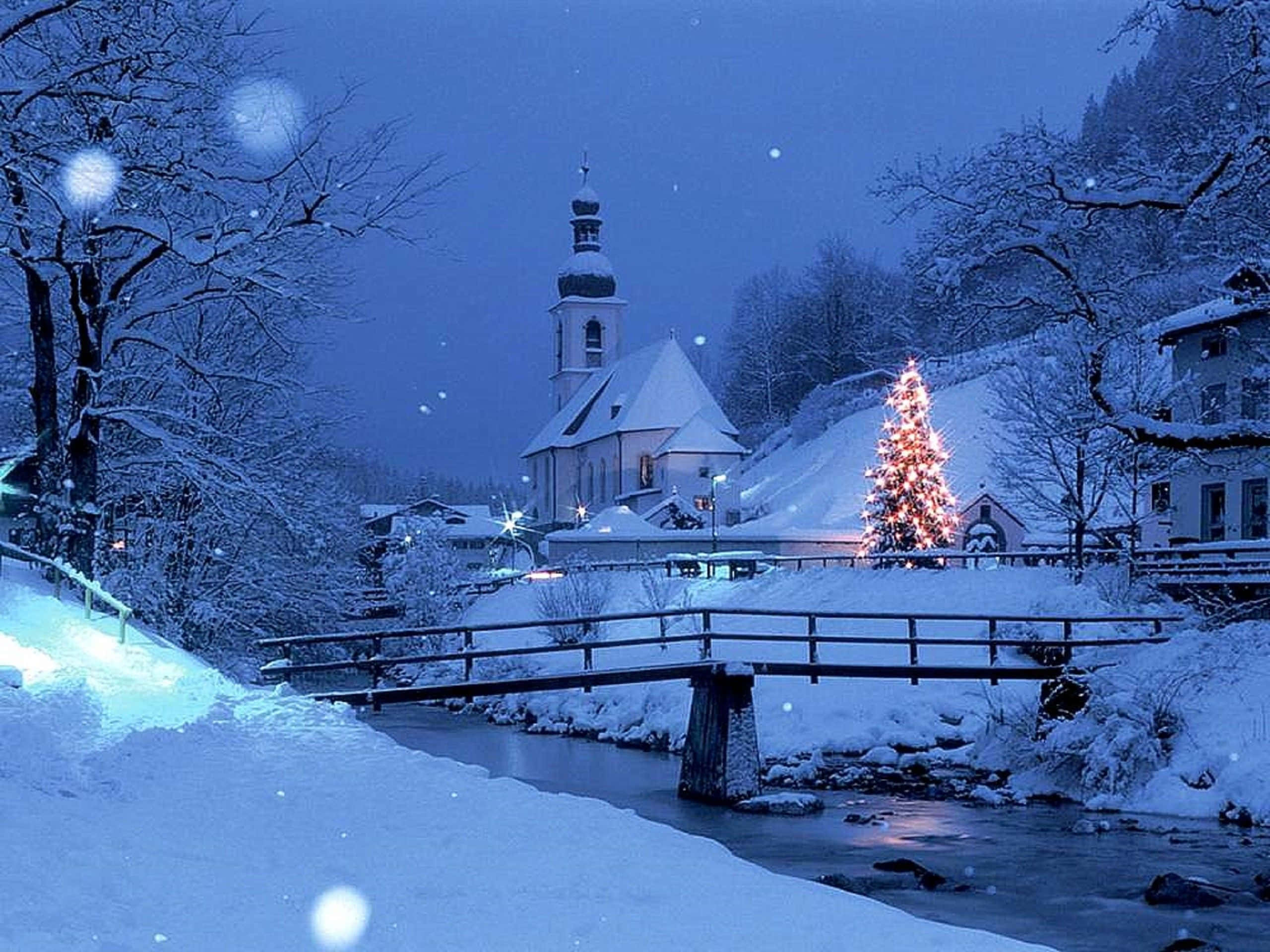 Snowy Village 桌布 壁紙 — 黃昏時分被雪覆蓋的村莊，河上的橋旁有發光的聖誕樹，背景是教堂。