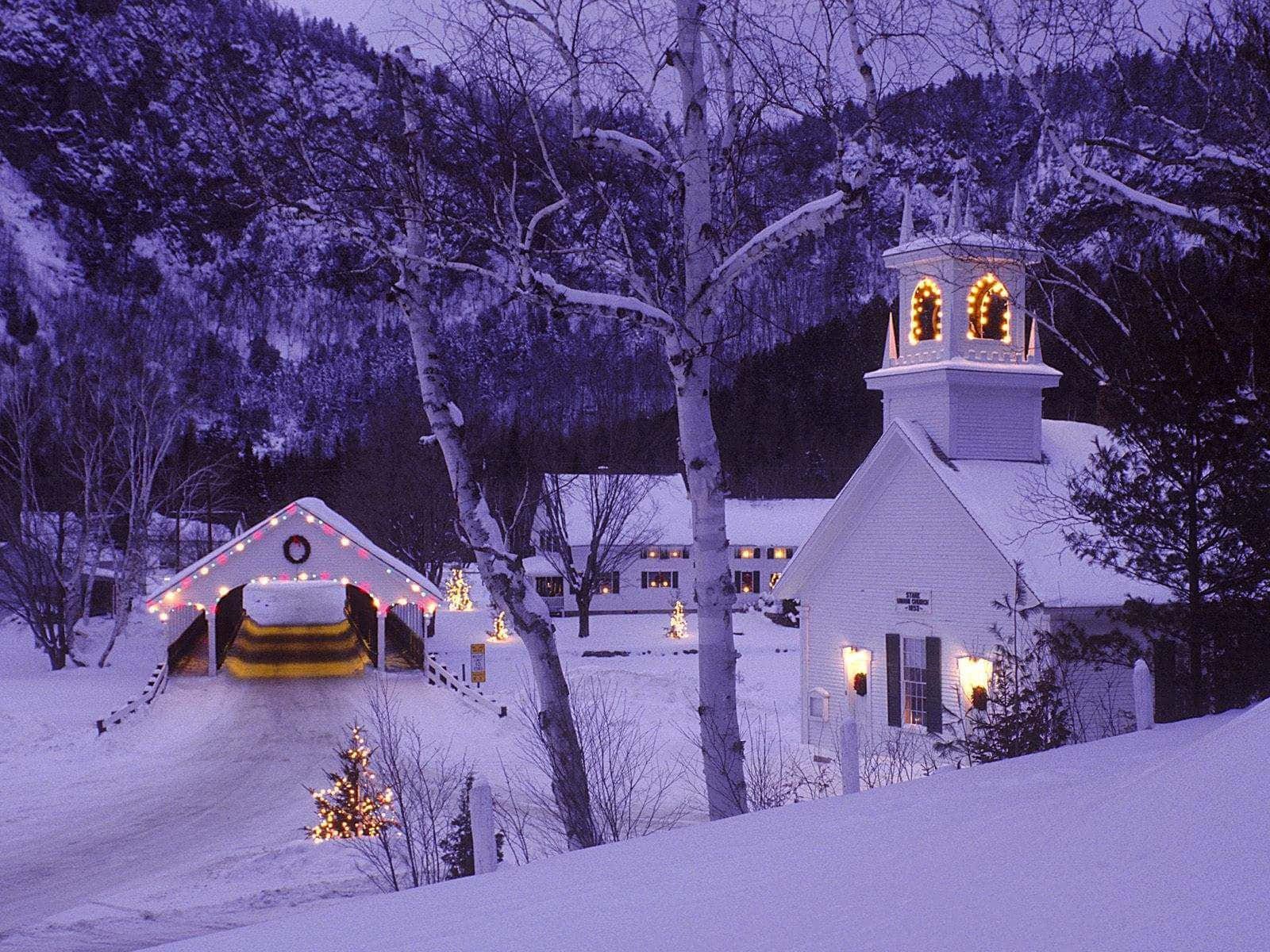 Winter Scene 桌布 壁紙 — 黃昏時分被雪覆蓋的村莊，燈火通明的教堂和有蓋橋樑在紫色山脈的映襯下溫暖地發光。