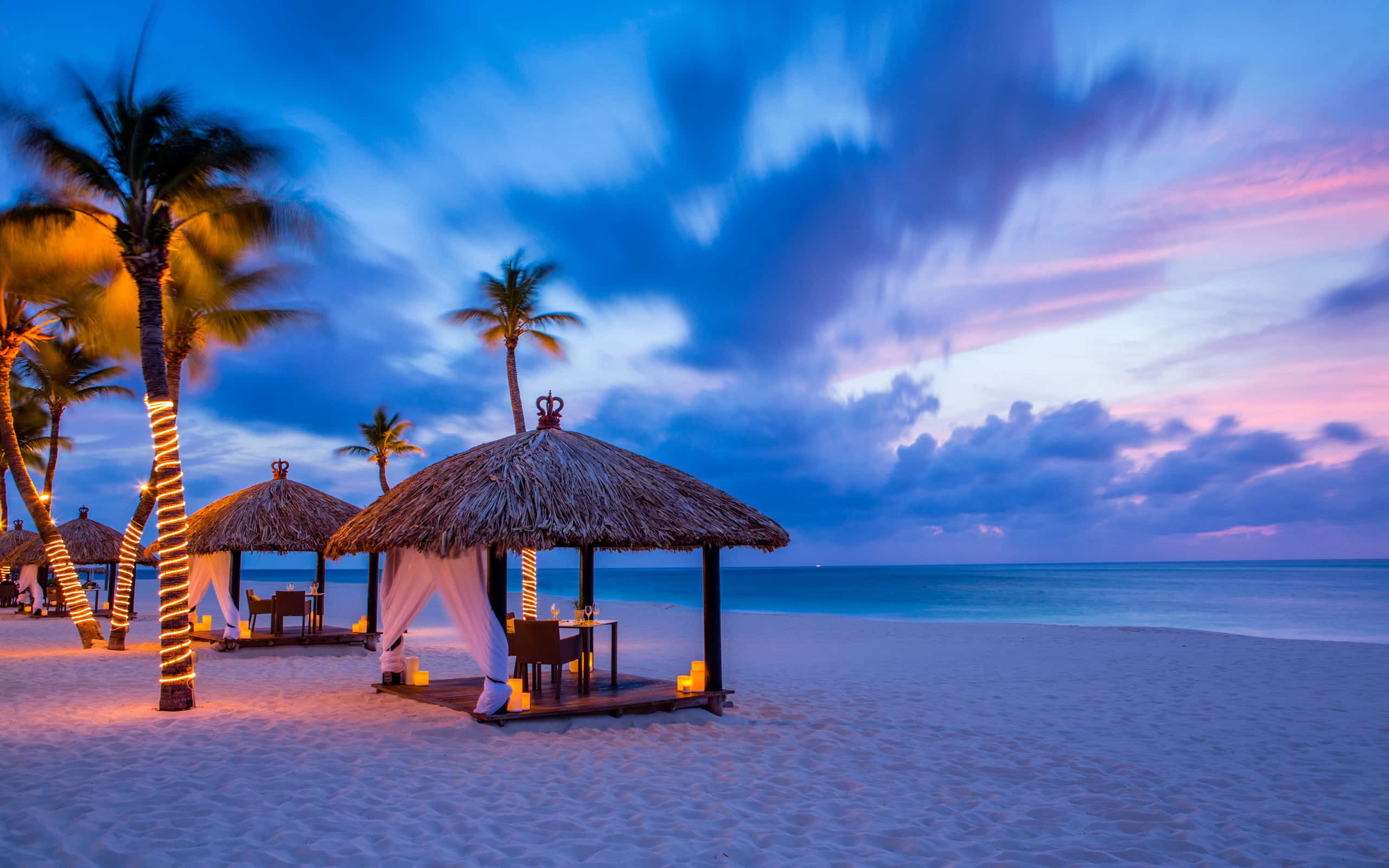 Beach Resort 桌布 壁紙 — 黃昏海灘場景，茅草屋頂涼亭在充滿活力的紫色和粉紅色天空下被蠟燭照亮。