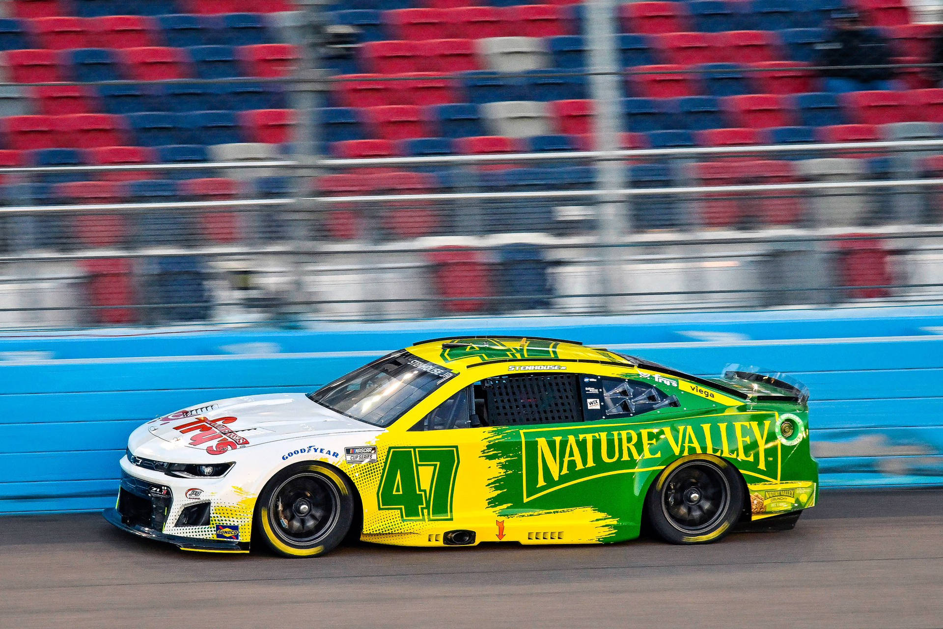一輛綠色和黃色的NASCAR賽車在賽道上疾馳，背景是模糊的體育場座位。