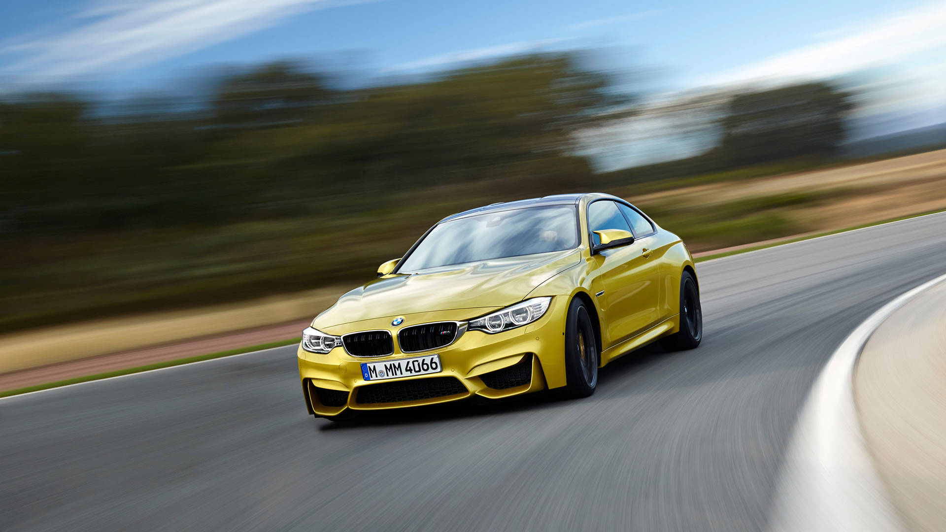 黃色BMW M車高速行駛 壁紙 — 一輛鮮黃色BMW M4 Coupe在彎曲的道路上疾馳，背景是模糊的綠樹和天空。