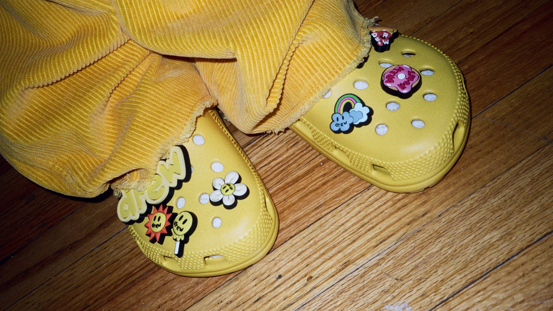 黃色Crocs配Jibbitz飾品 壁紙 — 黃色Crocs鞋綴有彩色飾物，放在木地板上，下方是黃色燈芯絨褲。