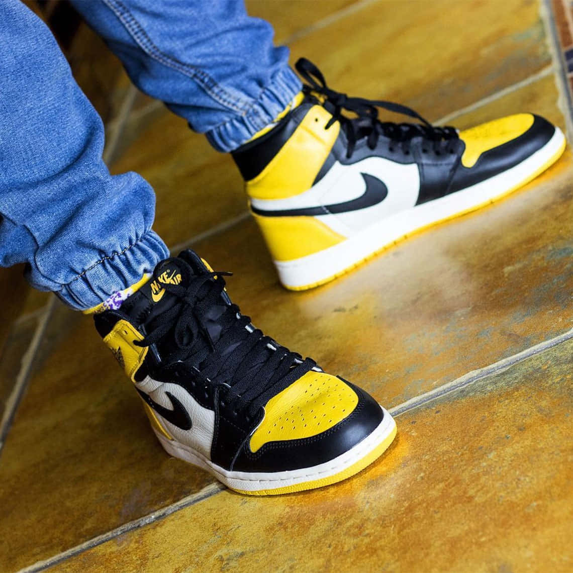 瓷磚地板上，穿著牛仔褲（褲腳摺起）的黃、黑、白色Nike Air Jordan 1運動鞋的人。