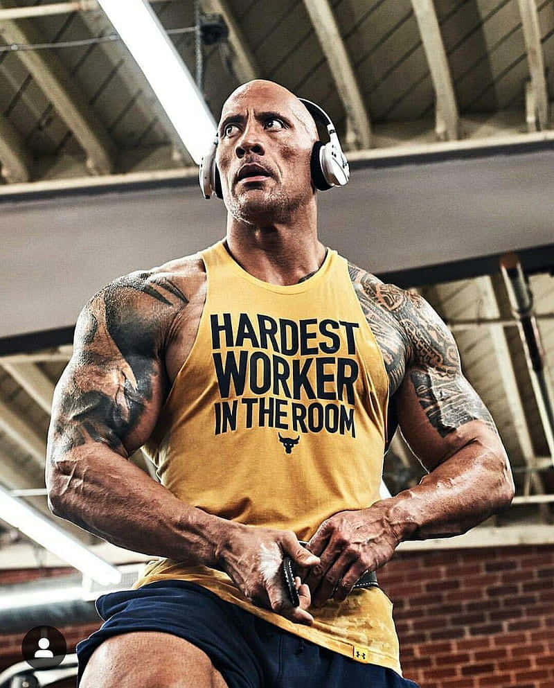 Dwayne Johnson 在健身房戴著耳機，身穿黃色背心舉重。