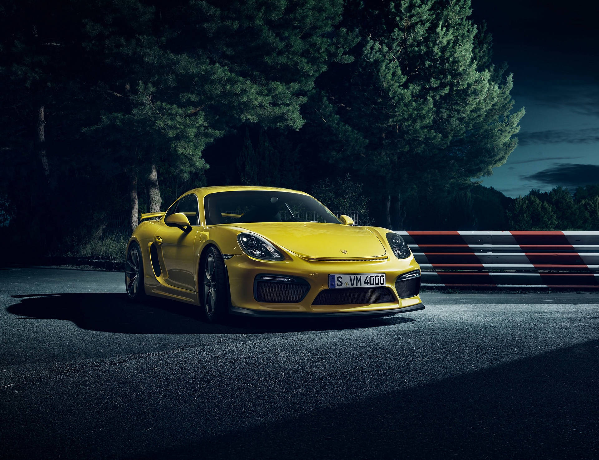 黃色保時捷Cayman夜景特寫 壁紙 — 一輛鮮黃色的保時捷 Cayman GT4 停在夜間賽道上，周圍有深色樹木和紅白相間的護欄。