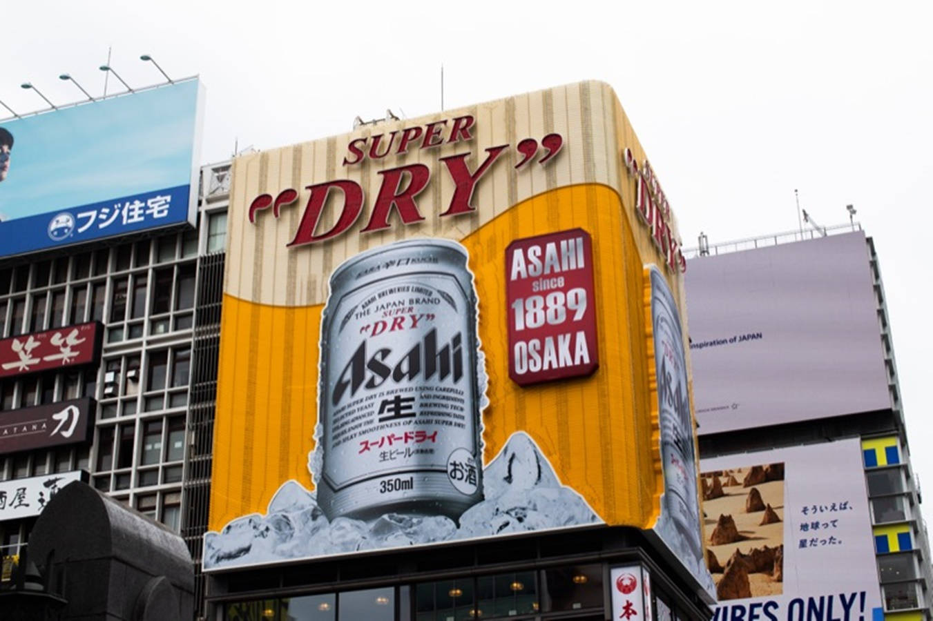 黃色啤酒廠朝日超乾 壁紙 — 一個巨大的Asahi Super Dry啤酒罐廣告，在陰天的天空下，主導了東京一座摩天大樓的外牆。