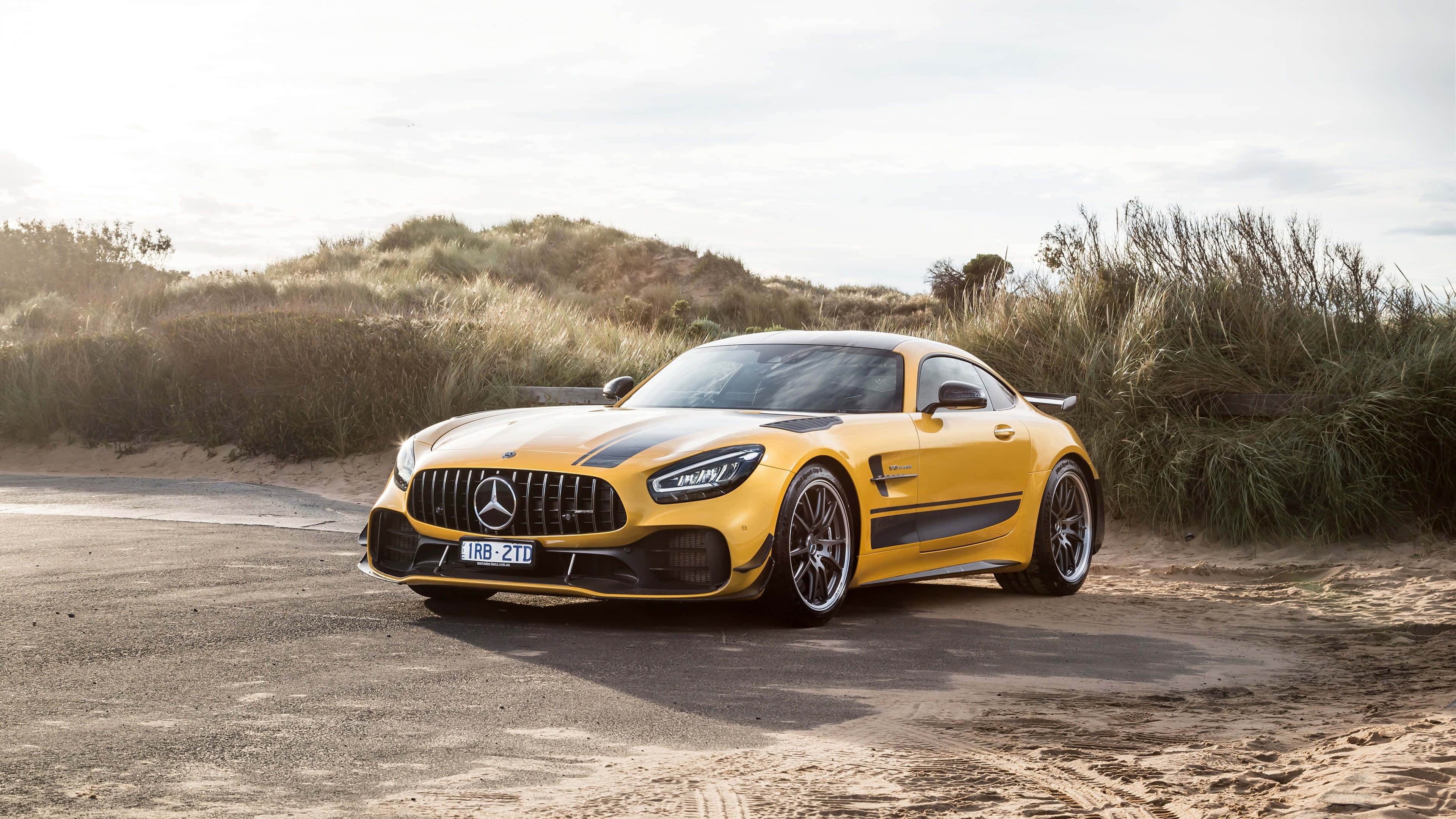 黃色梅賽德斯AMG GT R背景 壁紙 — 一輛鮮黃色的 Mercedes-AMG GT Black Series 跑車停在沙路上，旁邊有沙丘草，在柔和日光下。