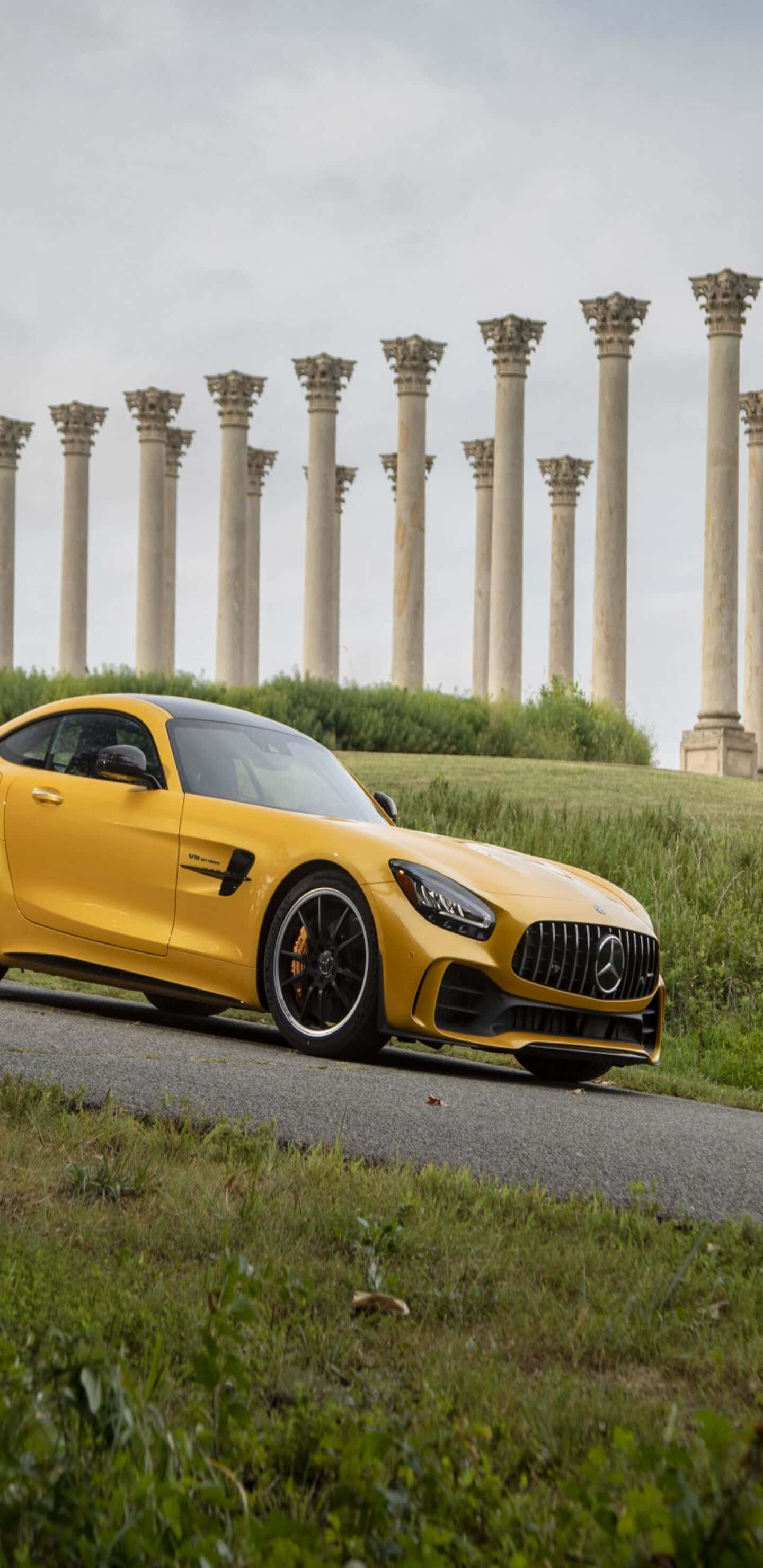 黃色梅賽德斯駛下山坡 Pixel 3XL AMG GT-R 背景 壁紙 — 一架鮮黃色嘅Mercedes-AMG GT跑車停泊喺一條路上面，前面係古老石柱，天空多雲。