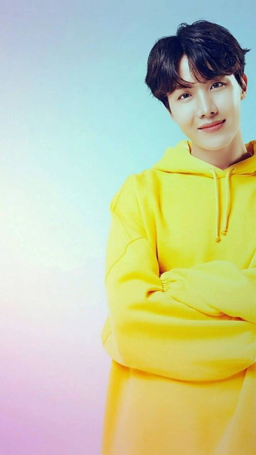 黃色毛衣的BTS J-Hope 壁紙 — 一位身穿亮黃色連帽衫的年輕男子自信地交叉雙臂，站在柔和漸變的天空背景前。