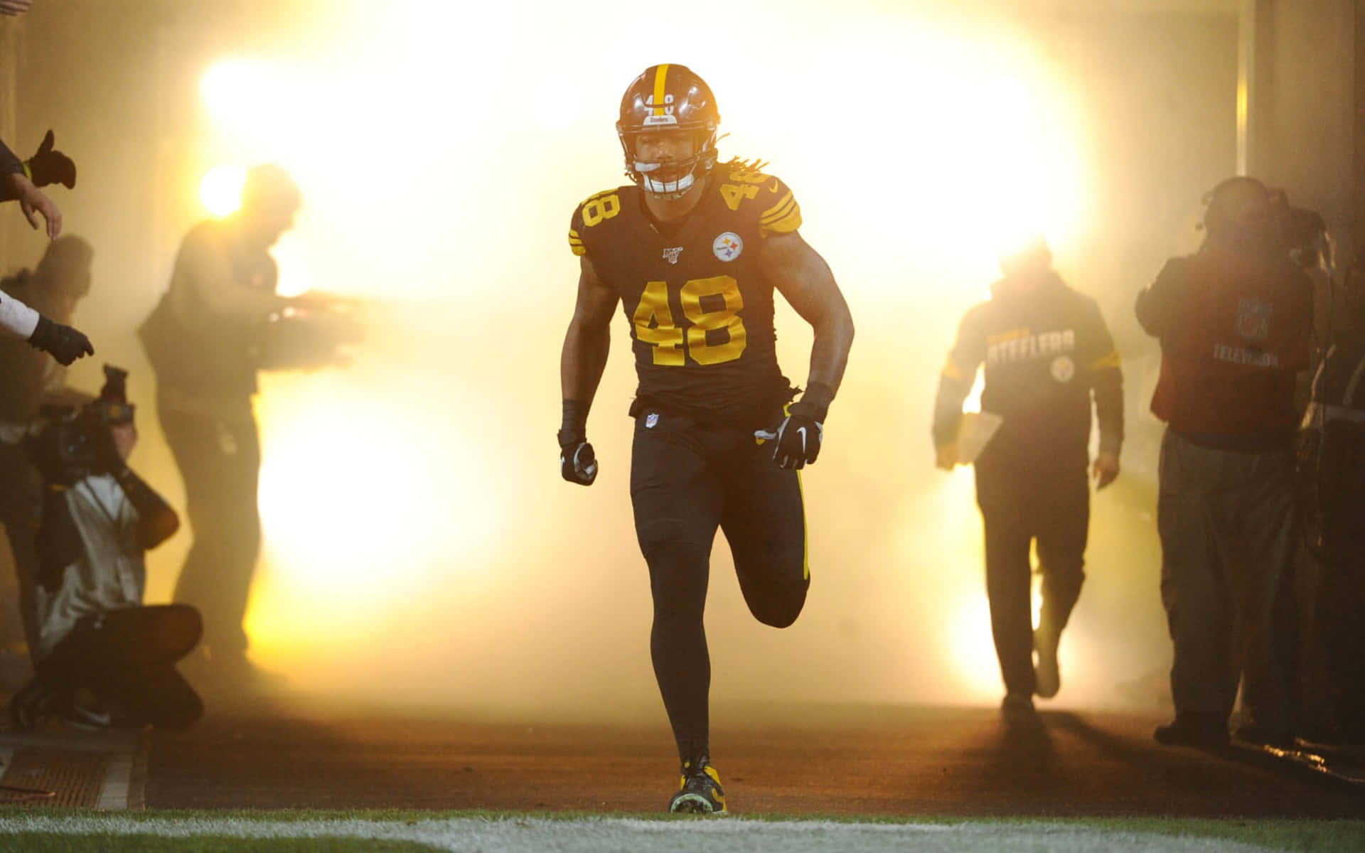 黃色燈光 Bud Dupree 壁紙 — 一名身穿黑金色球衣的匹茲堡鋼人隊美式足球員，在球場迷霧中奔跑，身後有明亮燈光。
