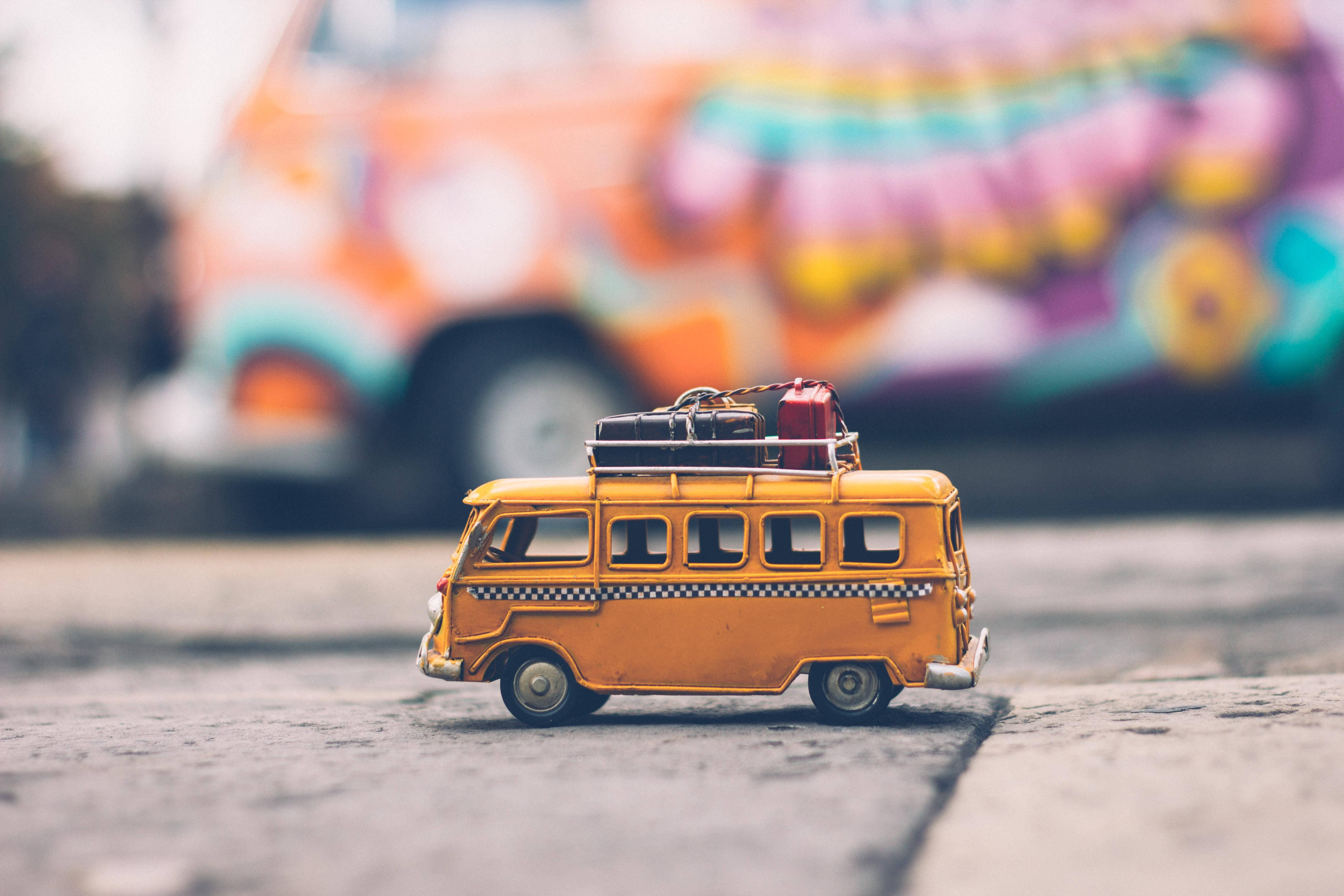 黃色玩具貨車度假 壁紙 — 一輛迷你橙色復古貨車連同行李放在人行道上，背景中有一輛色彩繽紛的塗鴉貨車模糊可見。