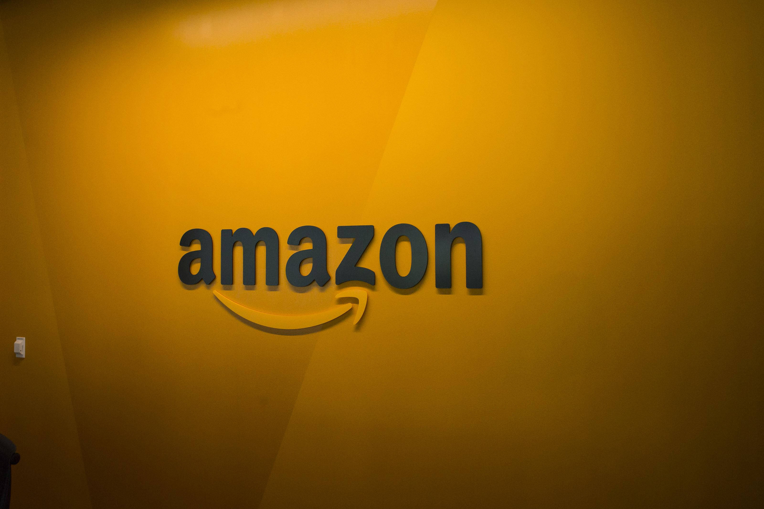 黃色背景上的Amazon標誌 壁紙 — 一個深灰色的簡潔亞馬遜標誌，帶有黃色箭頭，在溫暖的漸變黃色牆壁上微笑。
